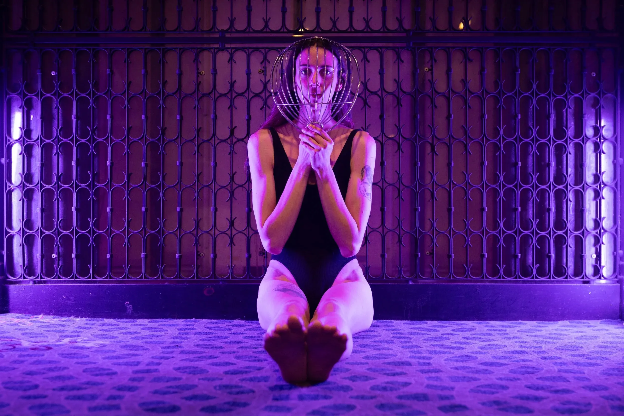 A woman sits beneath purple light before an ornate metal gate. She holds a thin metal cage in front of her face.