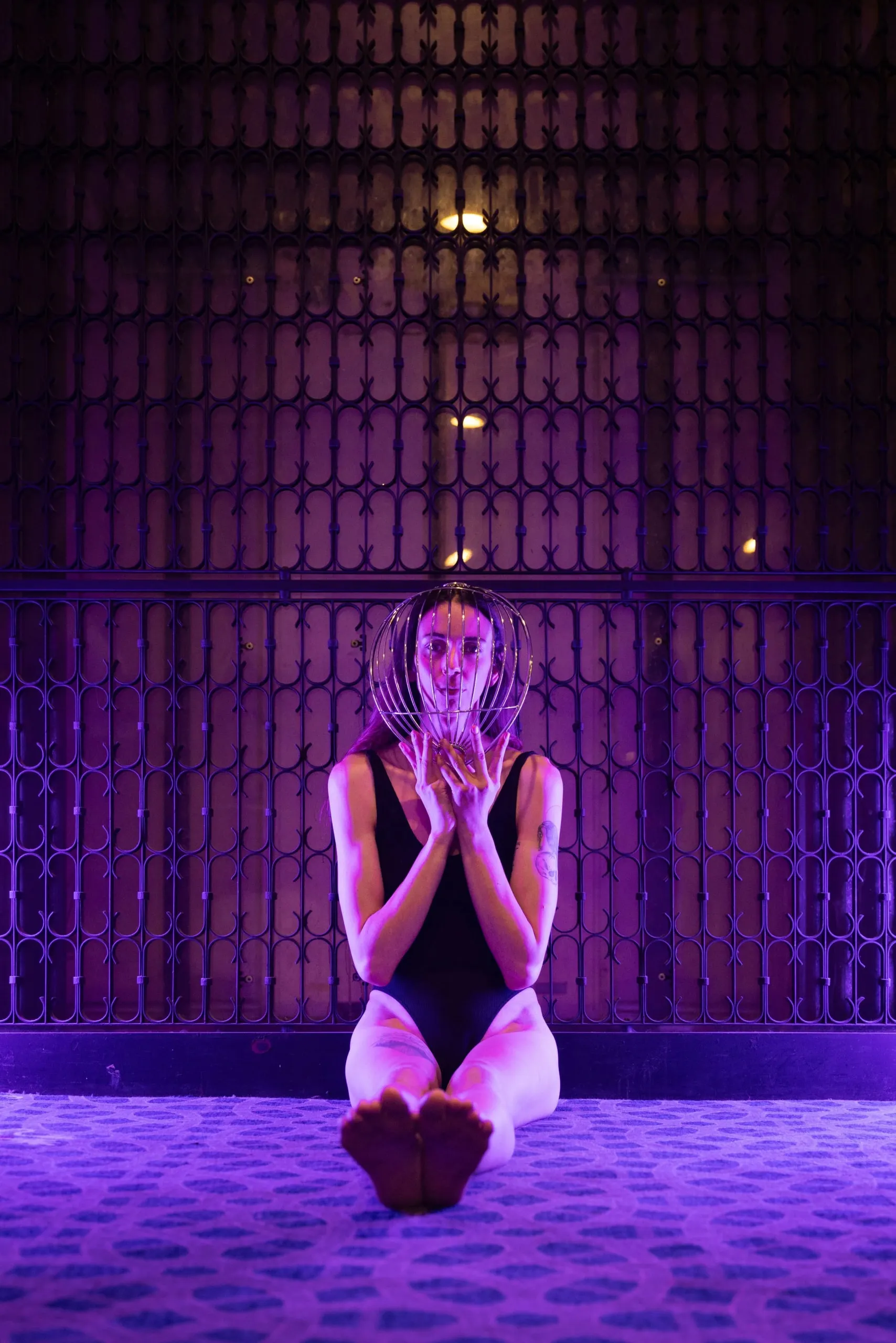 Lit in purple, a woman in a black bodysuit sits in front of an ornate metal gate, as she holds a thin metal cage in front of her face.
