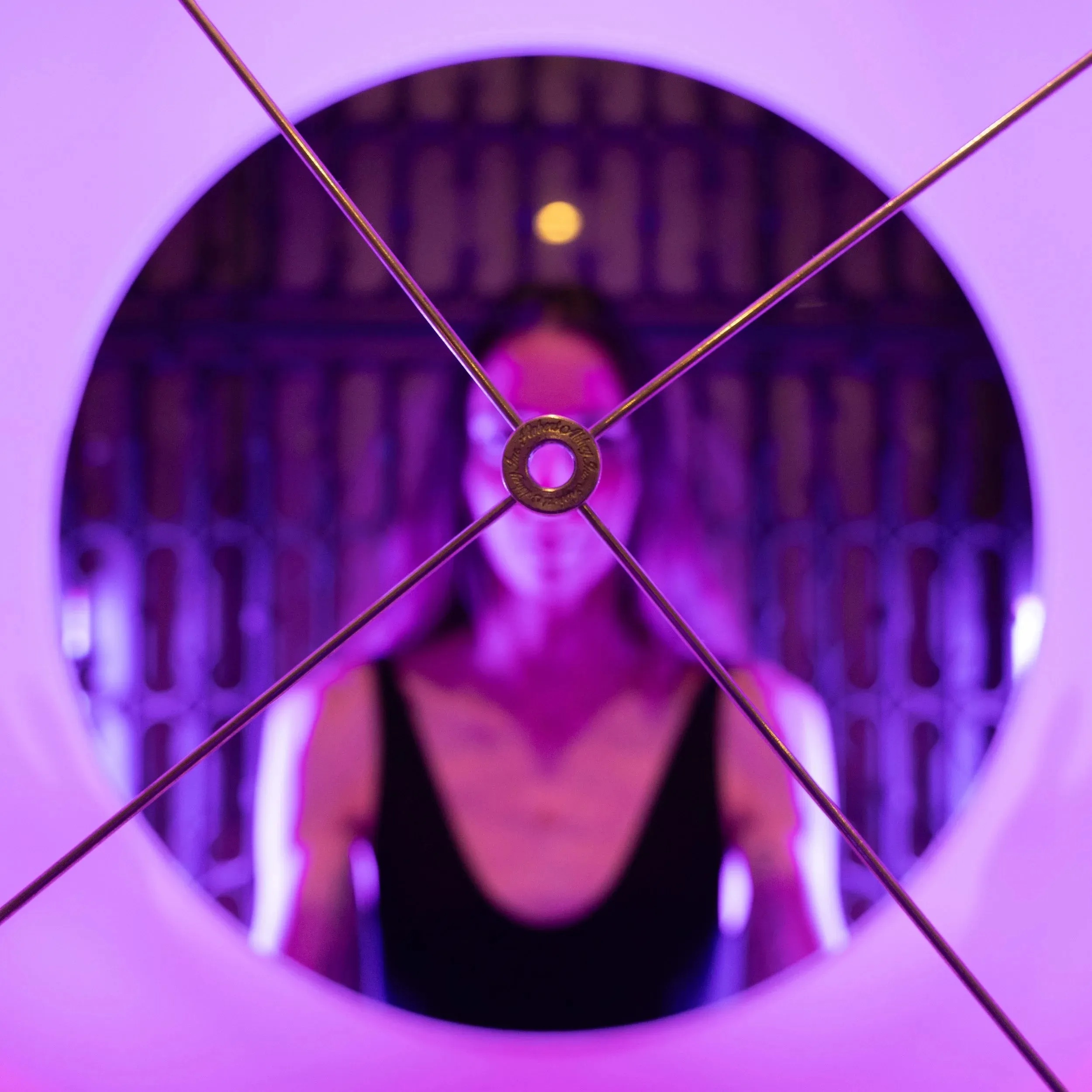 A woman peers through the circular opening of a purple-lit lampshade.