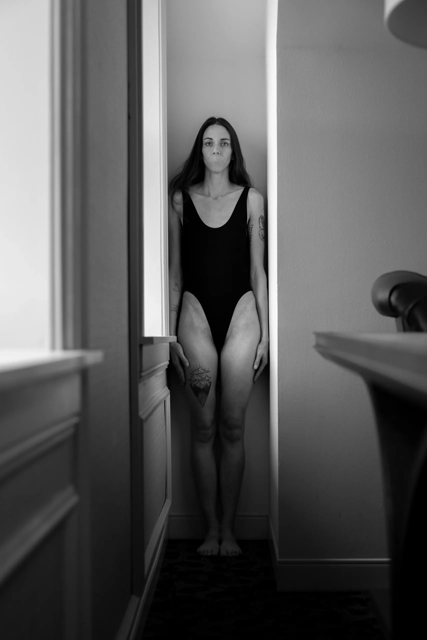 A surrealistic image shows a woman in black standing framed by a narrow nook in monochrome light; her mouth has been removed.