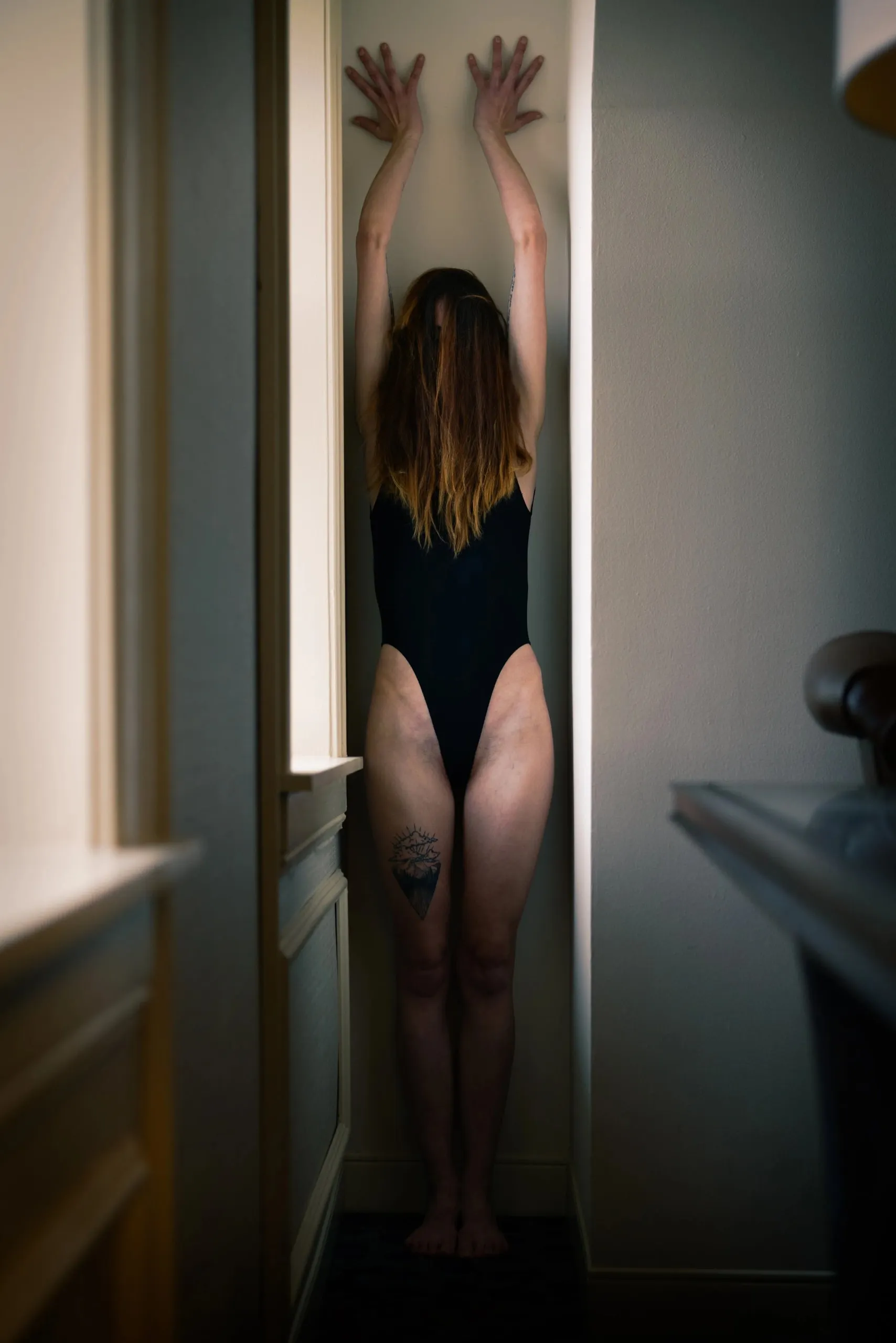 A woman reaches both hands overhead in a narrow sunlit recess, and her hair covers her face.