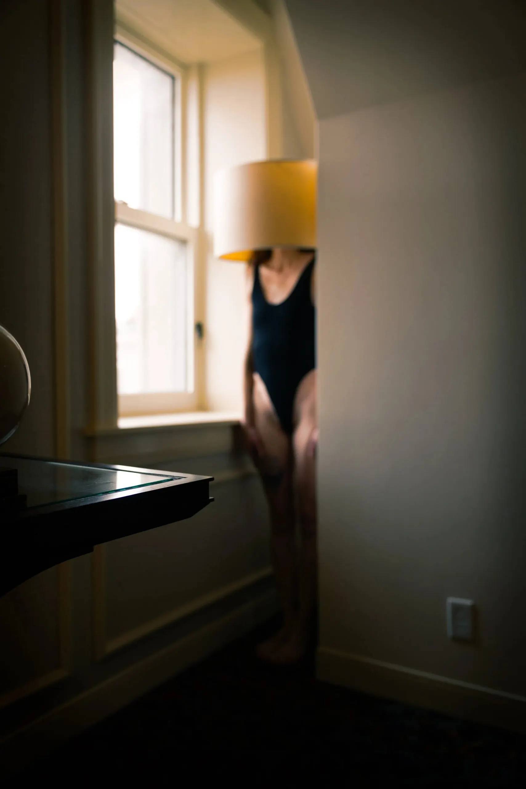 A woman stands next to a window, tucked away in a nook, with a lampshade on her head.
