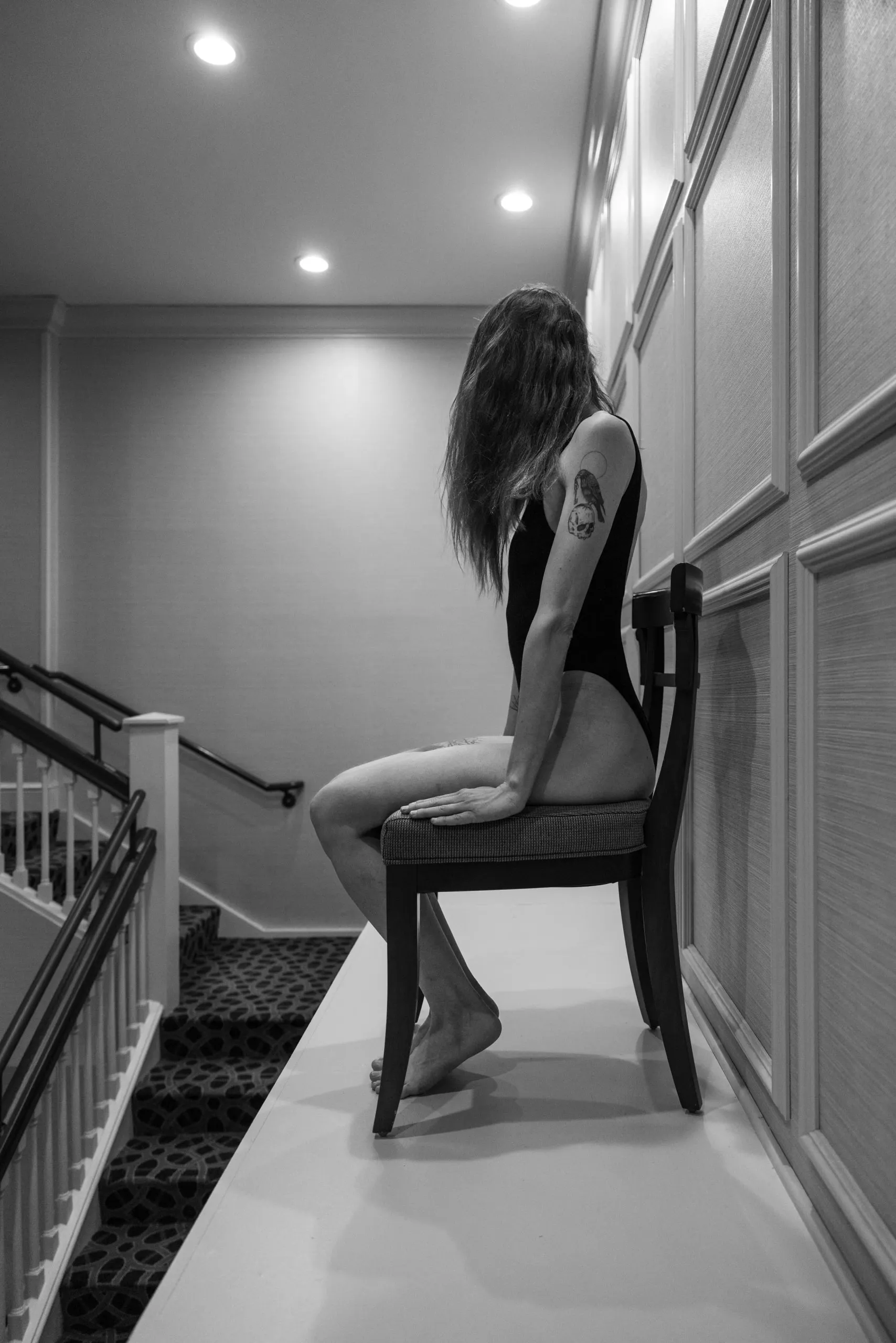 A woman in a black bodysuit sits on a chair, perched on a ledge above a flight of stairs.