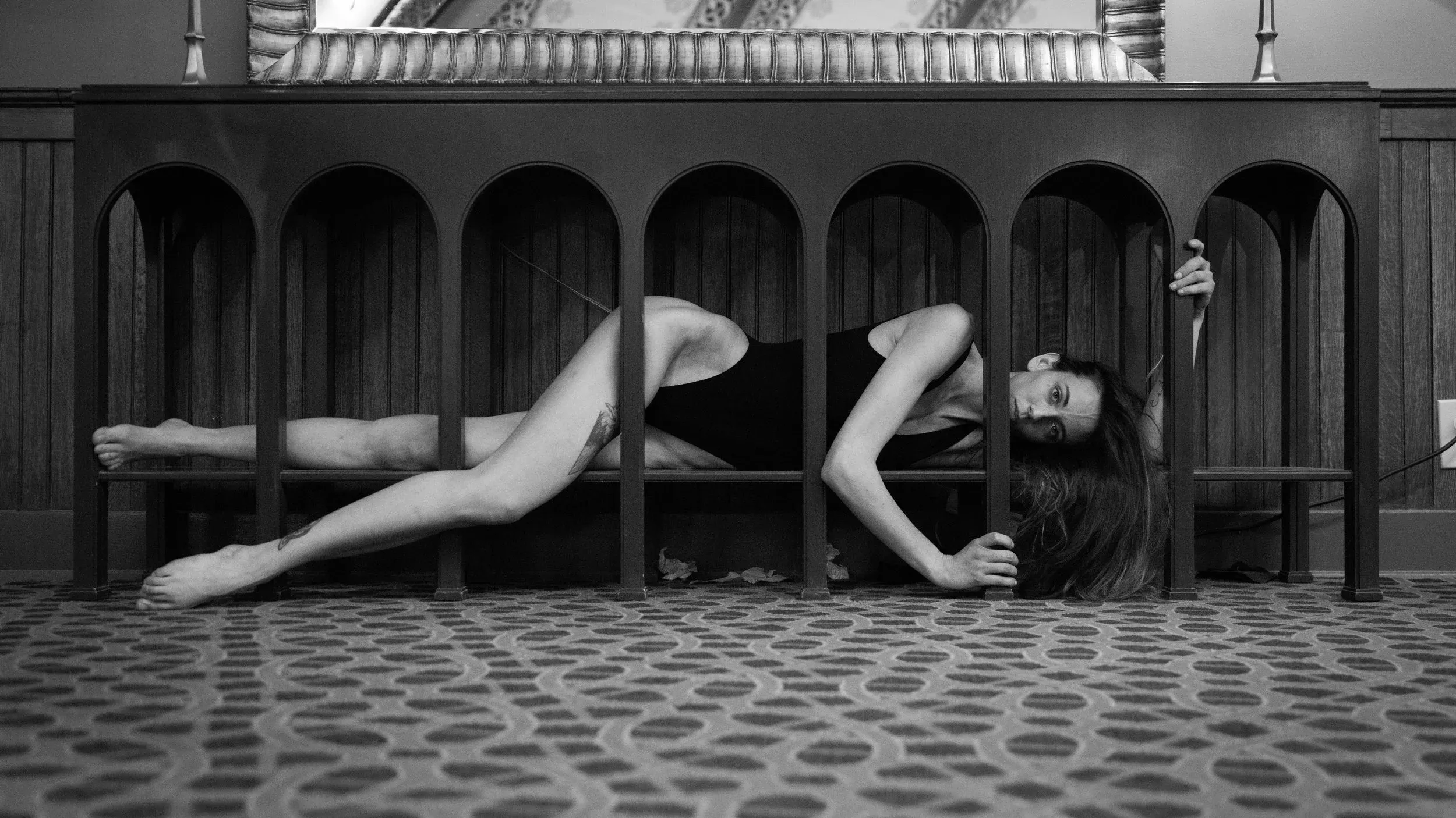 Fine art image of a woman in a black bodysuit entwined within many legs of a long narrow table.