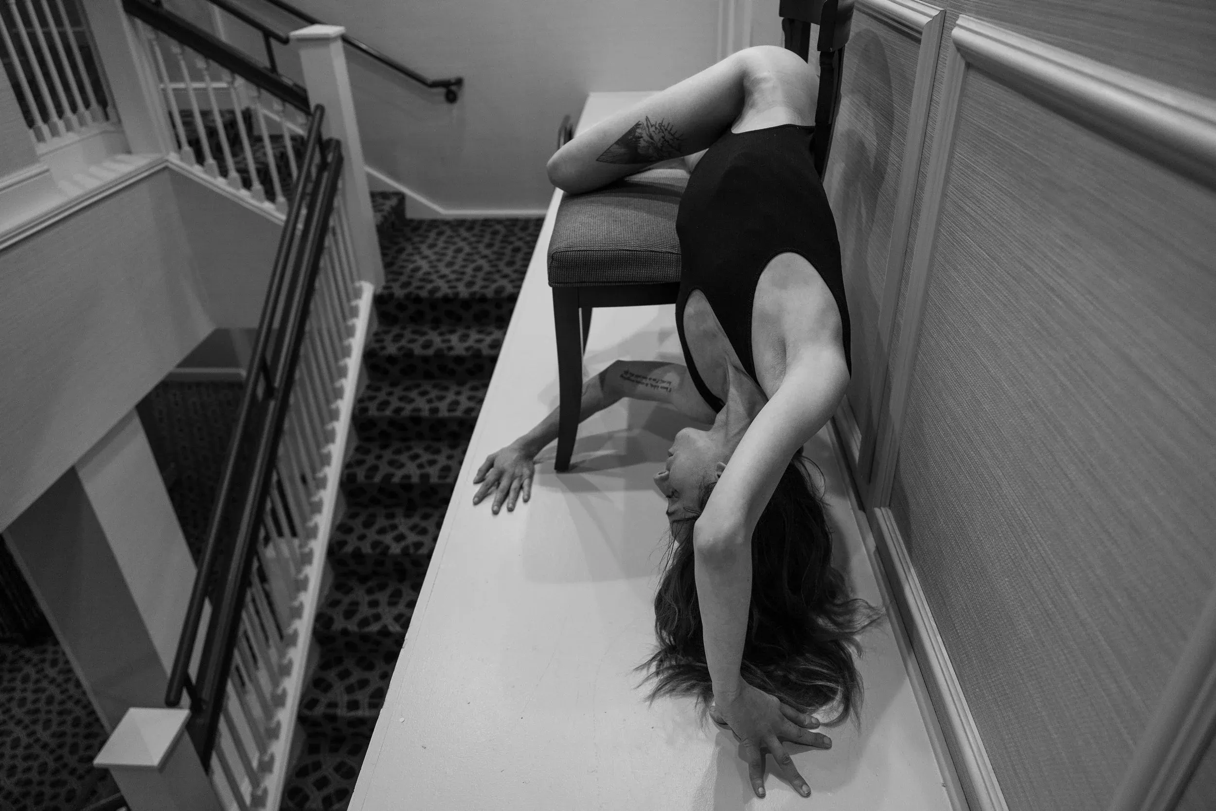 A tilted black-and-white view follows a woman stretching along a chair perched on a ledge above a flight of stairs.