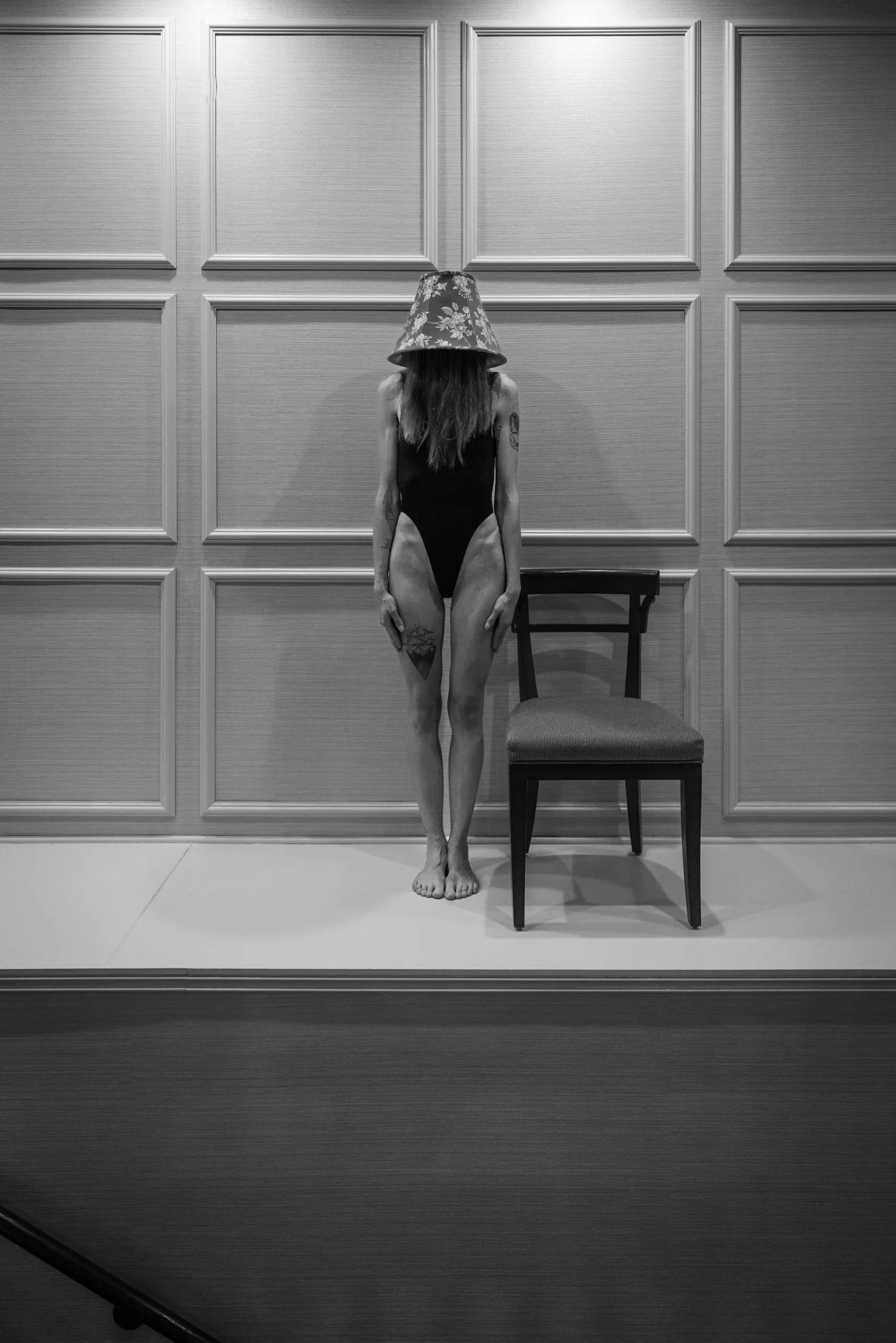 A woman wearing a black bodysuit stands at attention on a ledge over a flight of stairs, a lamp on her head.