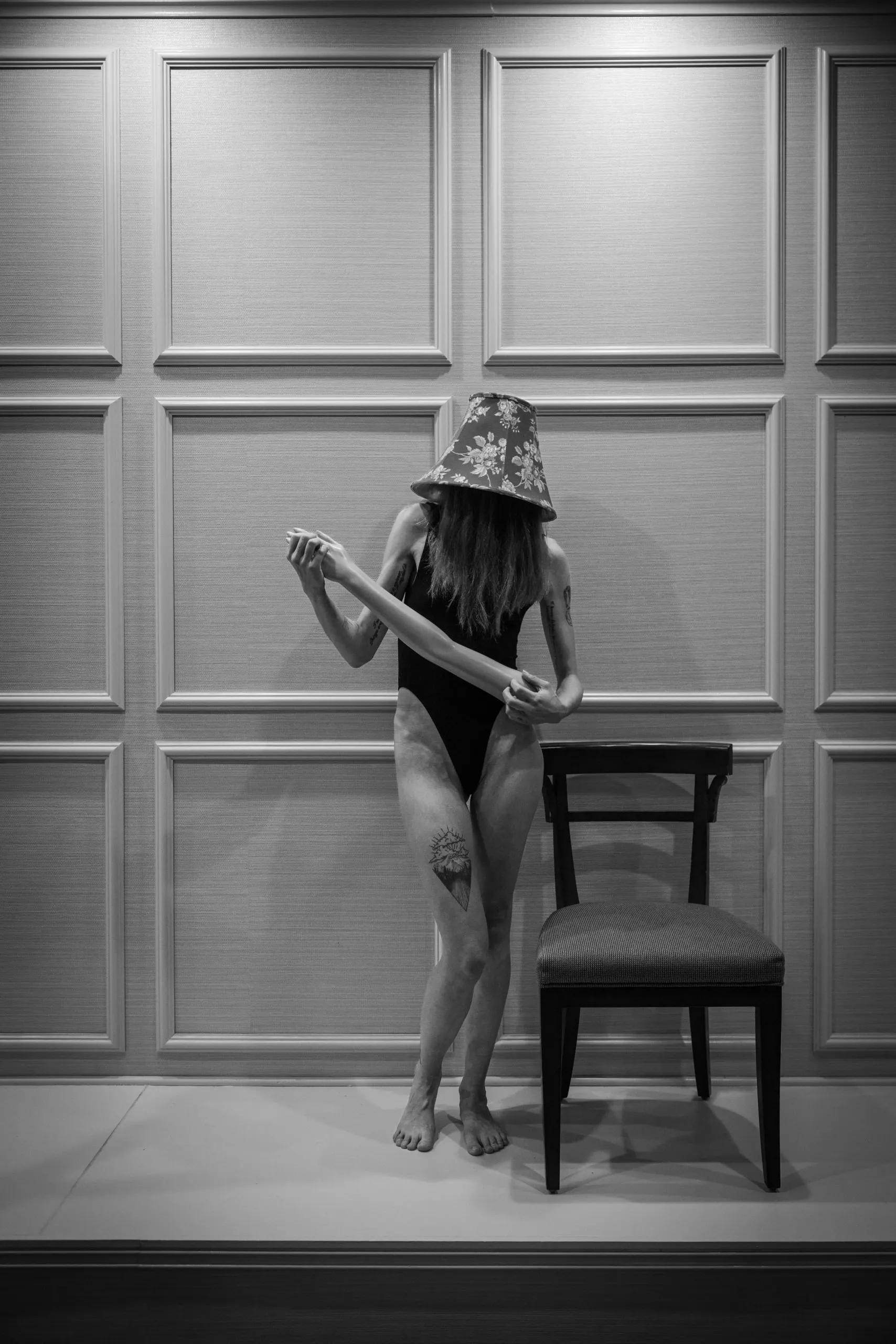 A woman wearing a bodysuit stands next to an empty chair, a lampshade on her head, as she plays air guitar with a mannequin arm.