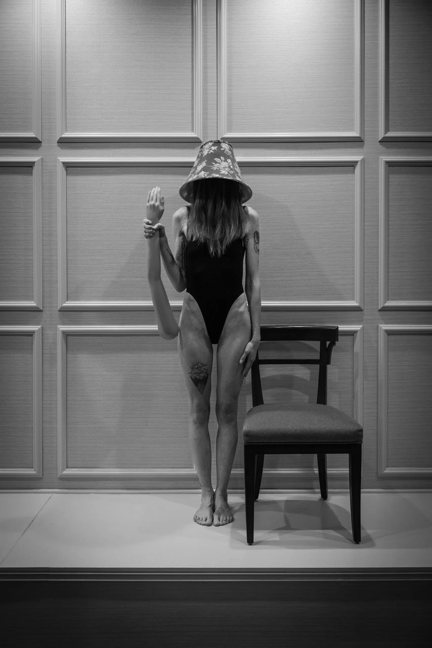 A woman in a black bodysuit stands next to an empty chair with a lampshade on her head and a mannequin arm in her right hand.