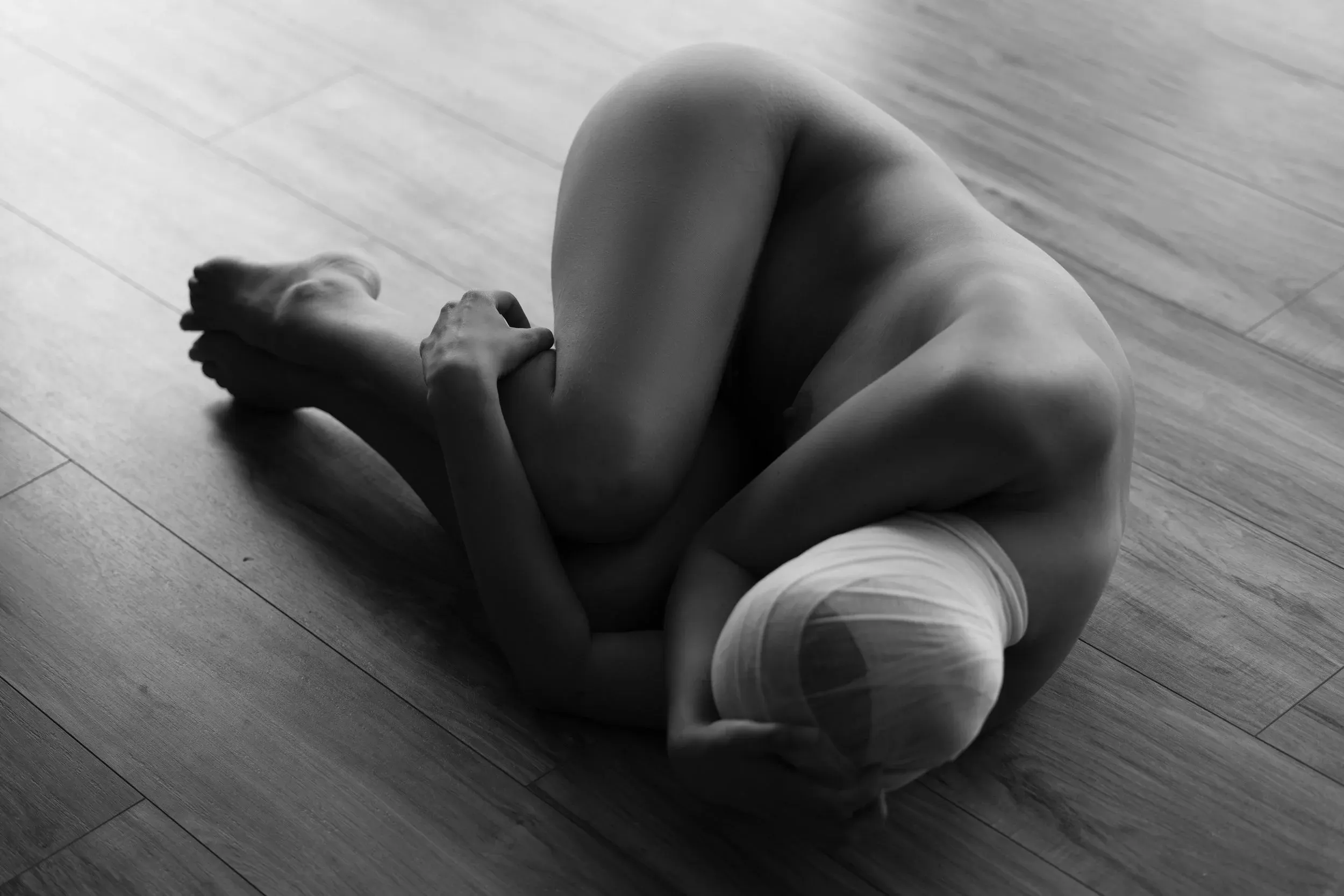 A nude woman lies curled on her side across a bright wood floor, her head wrapped in gauze.