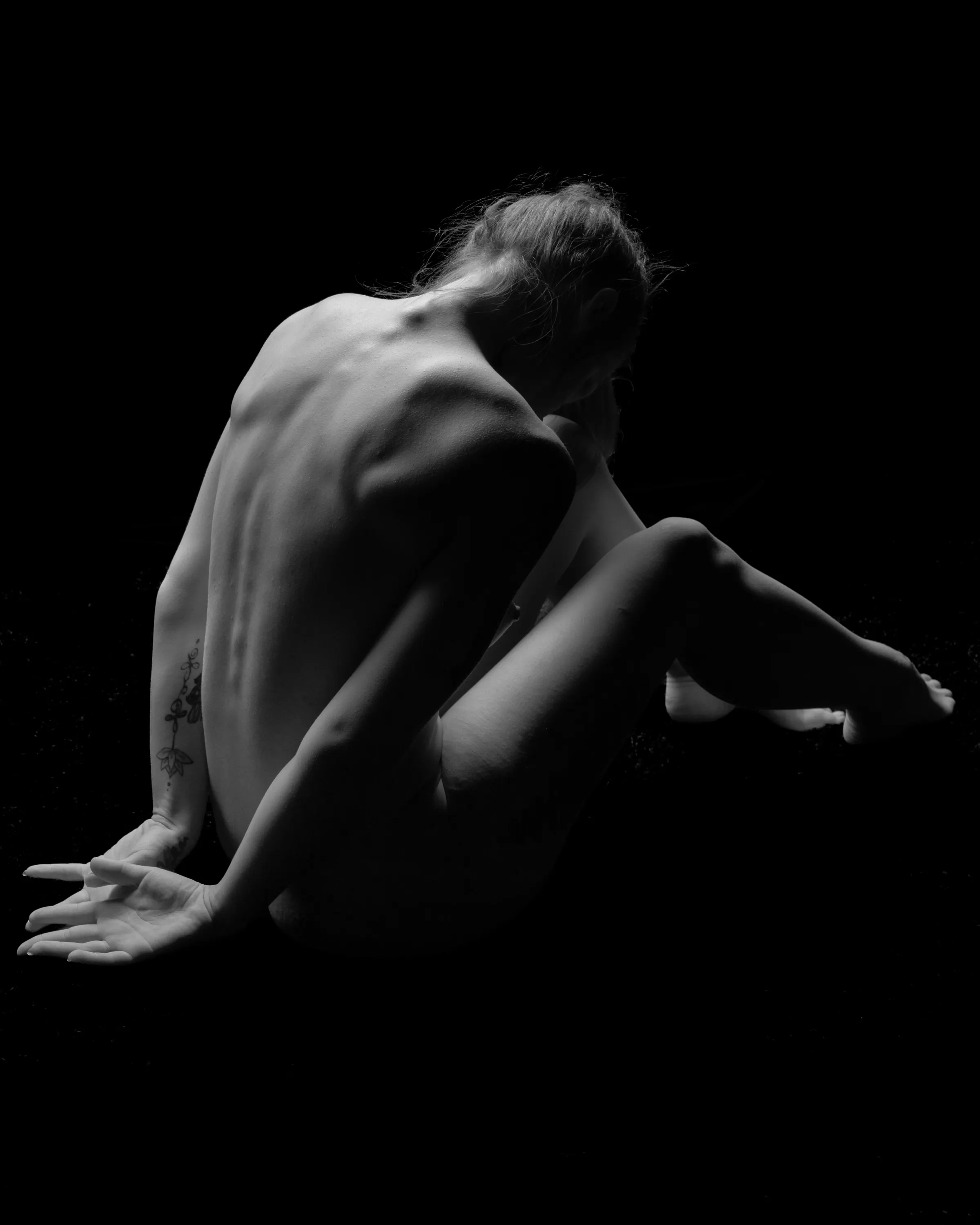 A black-and-white chiaroscuro image of a woman seated in a black void, facing away from the viewer with her back and legs visible, head bowed, and arms behind her back.