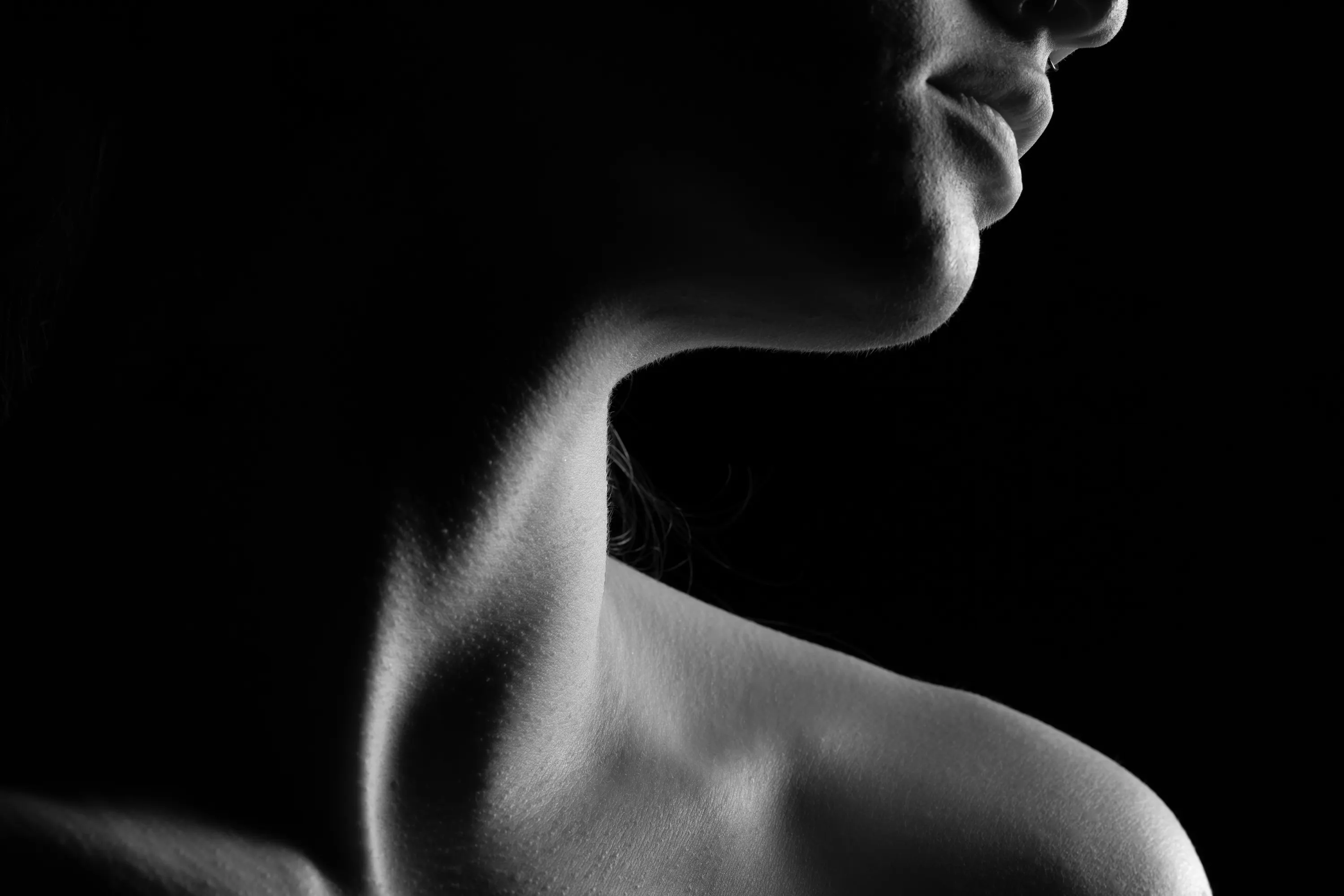 Close-up chiaroscuro image of a woman's shoulder, neck, chin, and mouth.