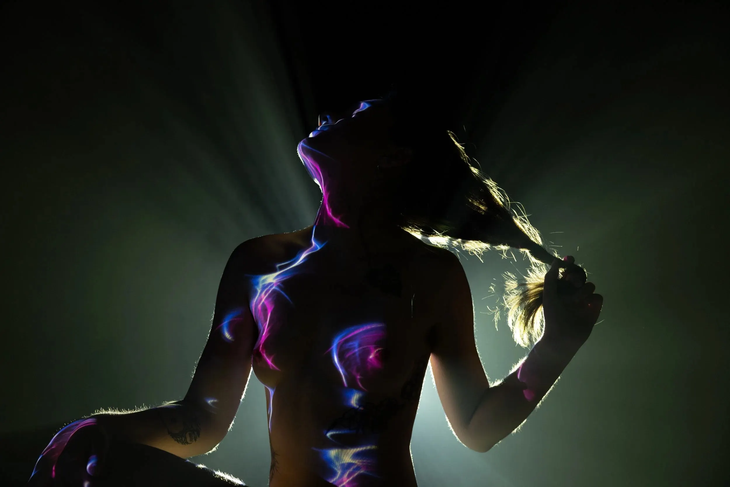 A woman pulls her hair as she sits, her body mostly a silhouette, lit from behind with subtle violet patterns projected onto her skin.