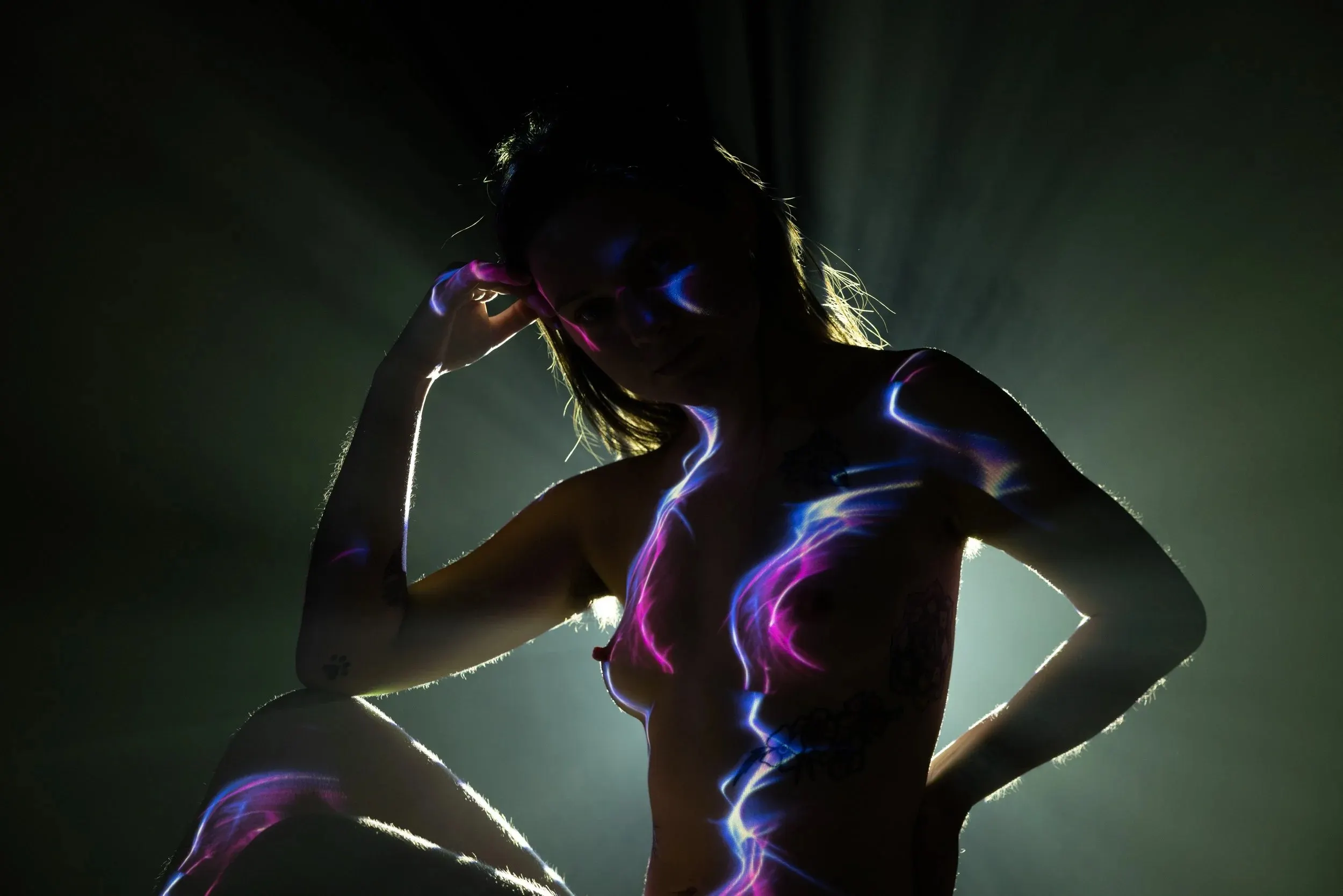 A backlit woman with subtle purple and blue patterns projected onto portions of her skin. The air is hazy, letting beams of light show.