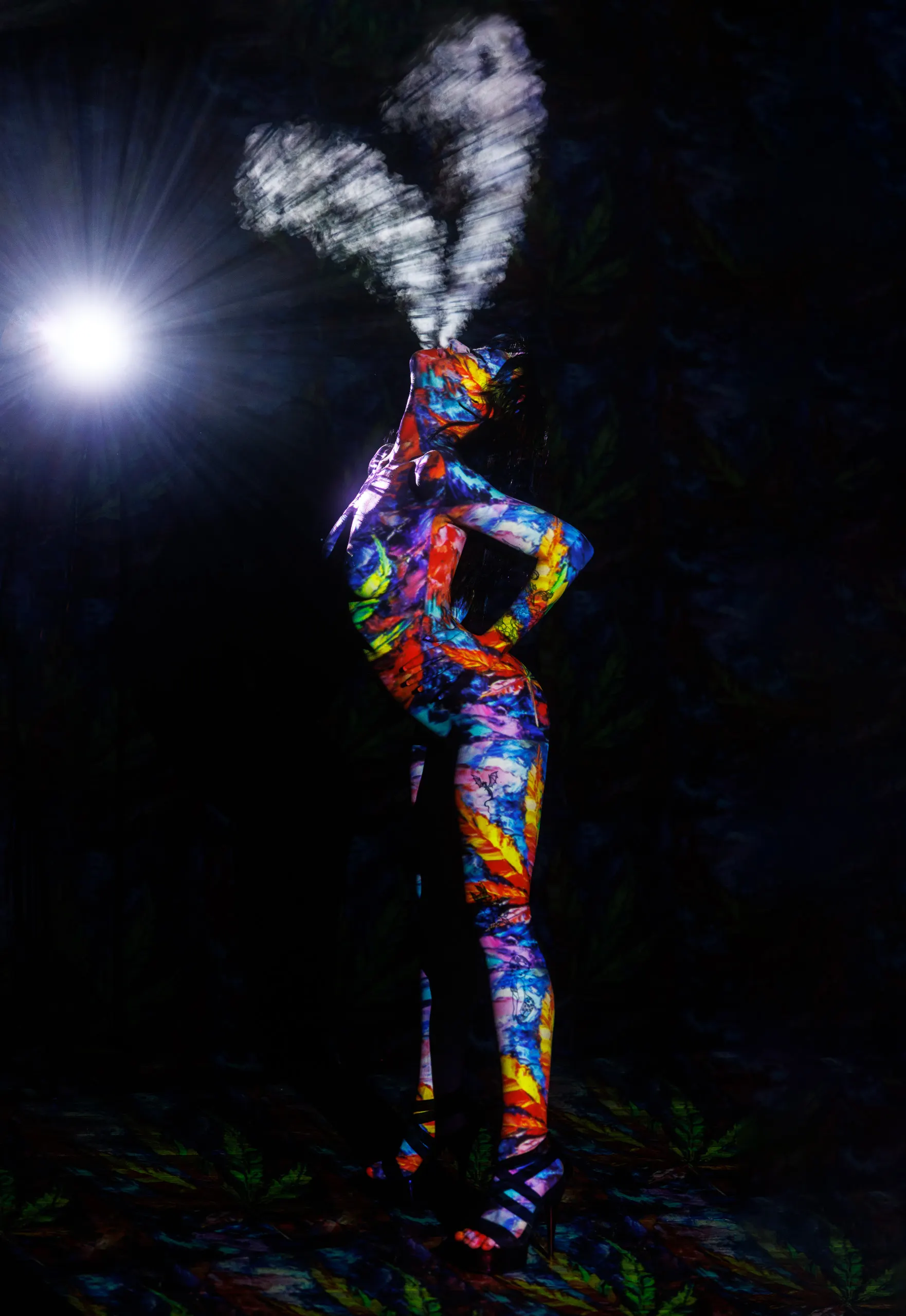 A woman stands in profile, hands on her hips, head tipped back as she exhales a plume of smoke split into two. Multicolored abstract patterns are projected across her body against a dark background. A beam of light from a projector is pointed toward the viewer.