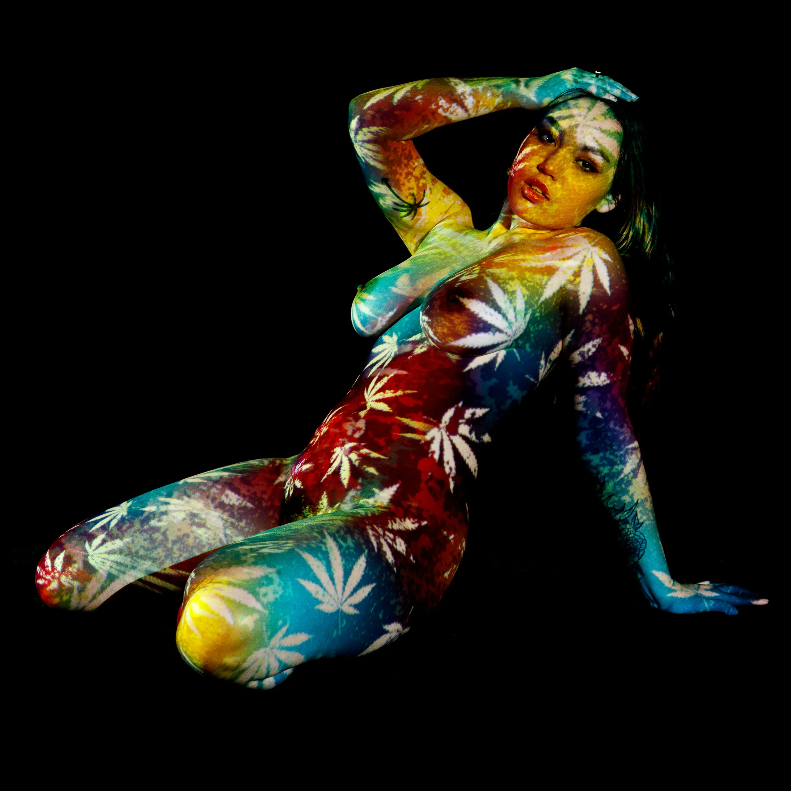 A woman reclines on one hip with an arm raised behind her head, white cannabis leaves projected over broad bands of red, yellow, and blue light across her whole body.
