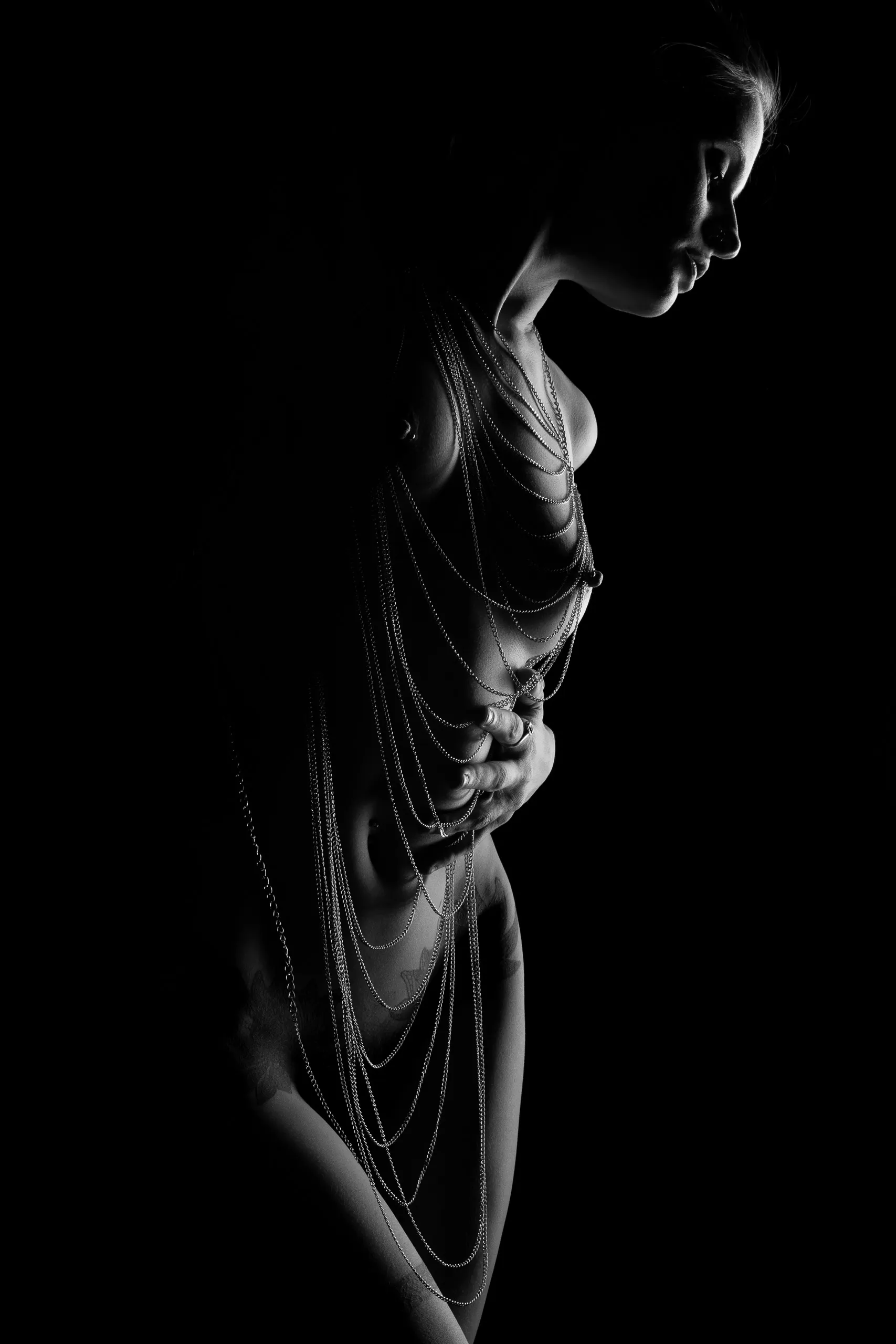 A woman stands in profile, fine silver chains draped from her shoulder down across her torso. She gathers the strands at her waist with one hand, lit in dramatic black-and-white side light.