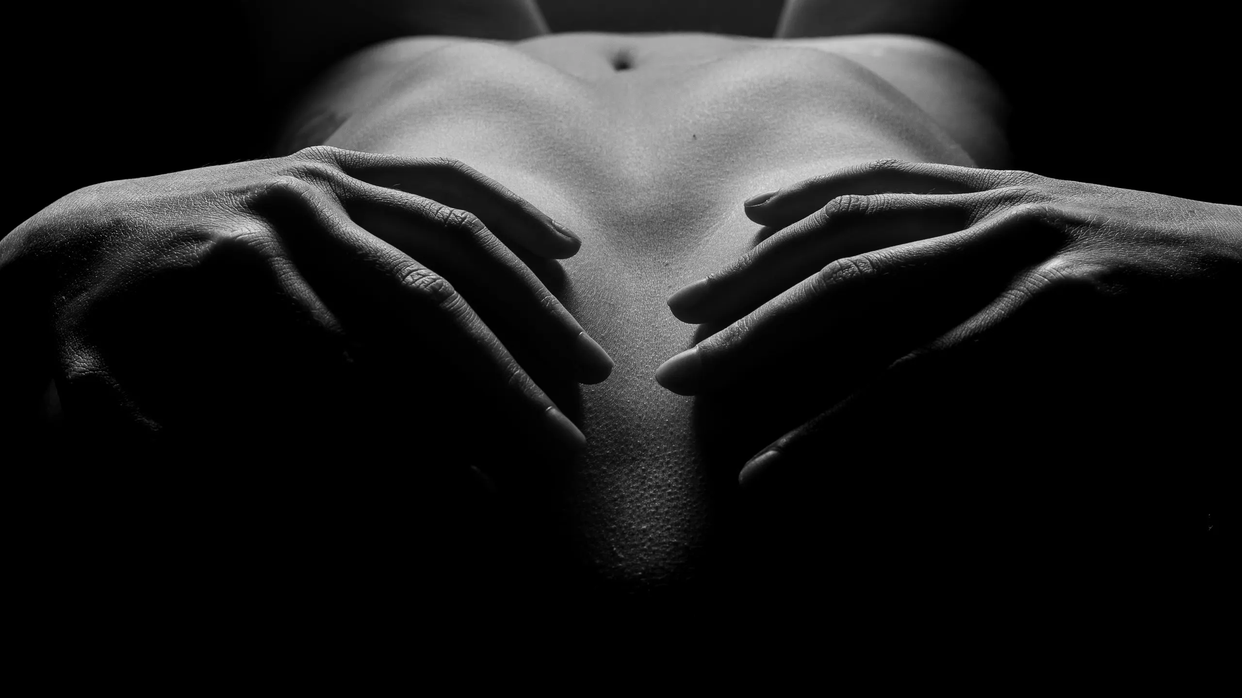 A close, low-light view of two hands resting on breasts, fingers spread and framing the navel in soft directional light.
