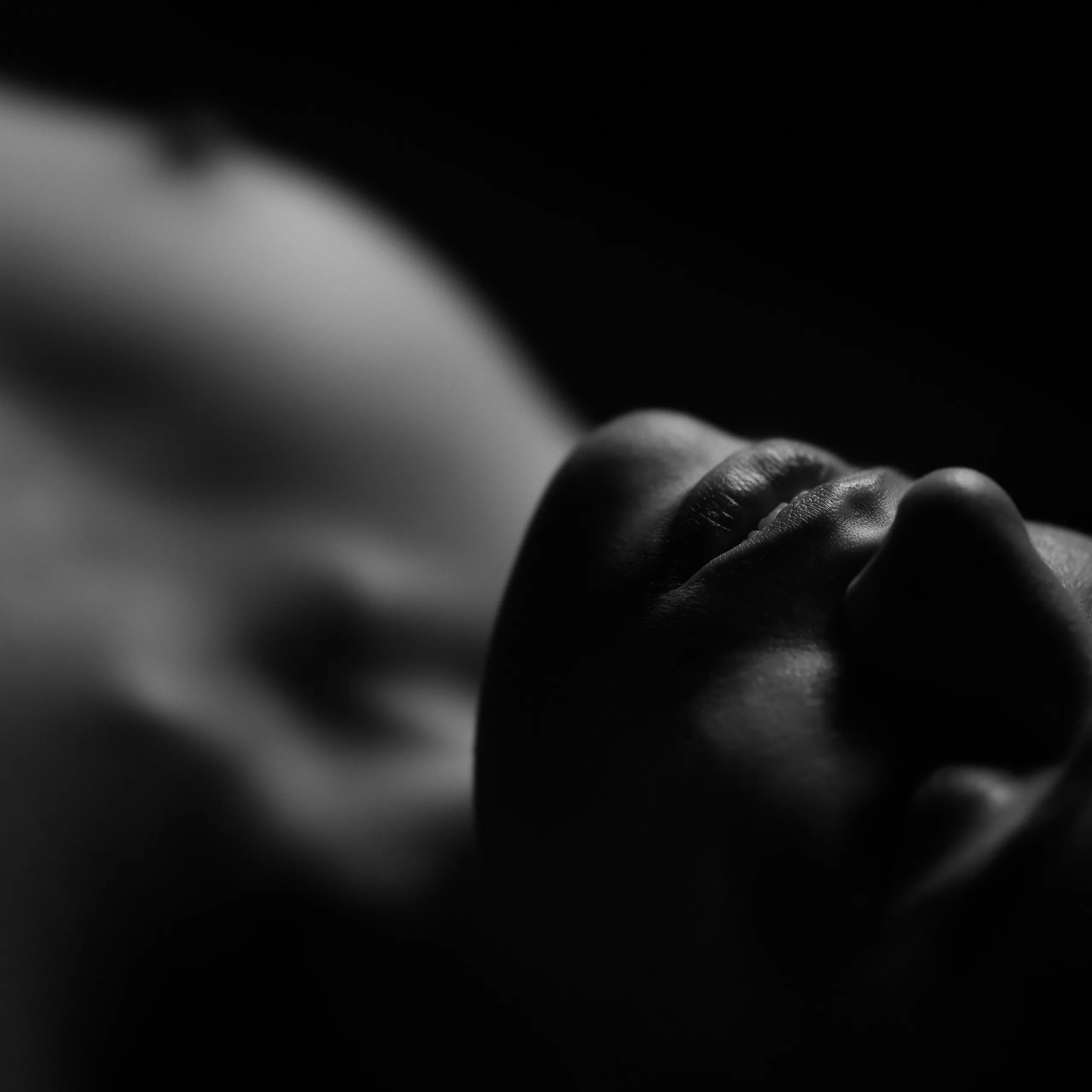 A tight, low-light macro focused on a woman's lips, soft focus blurring her body beyond it into pale shapes against black.