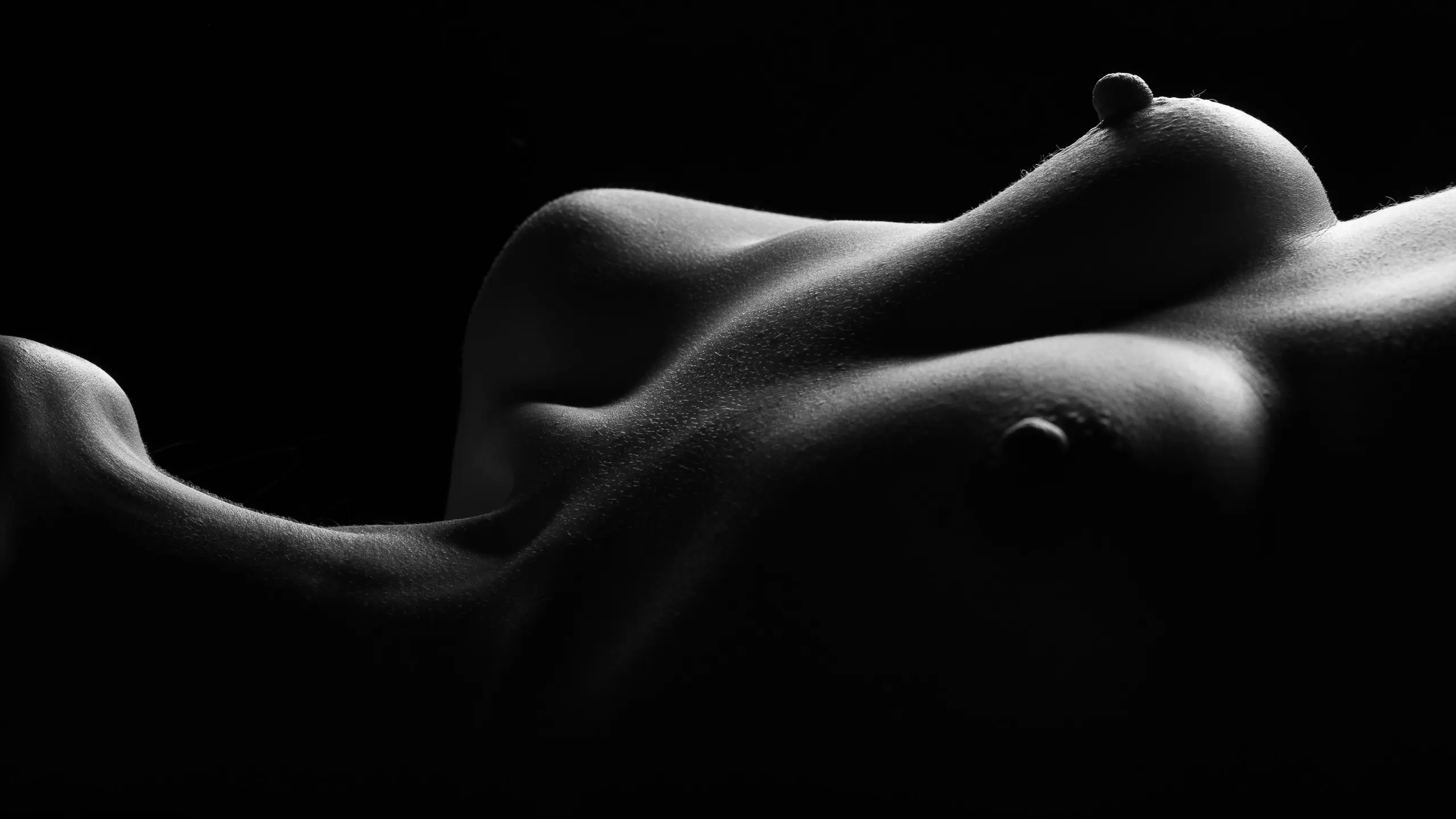 An extreme close study of overlapping curves where torso meets chest, rim-lit in white against black, a nipple catching the light at the edge of frame.