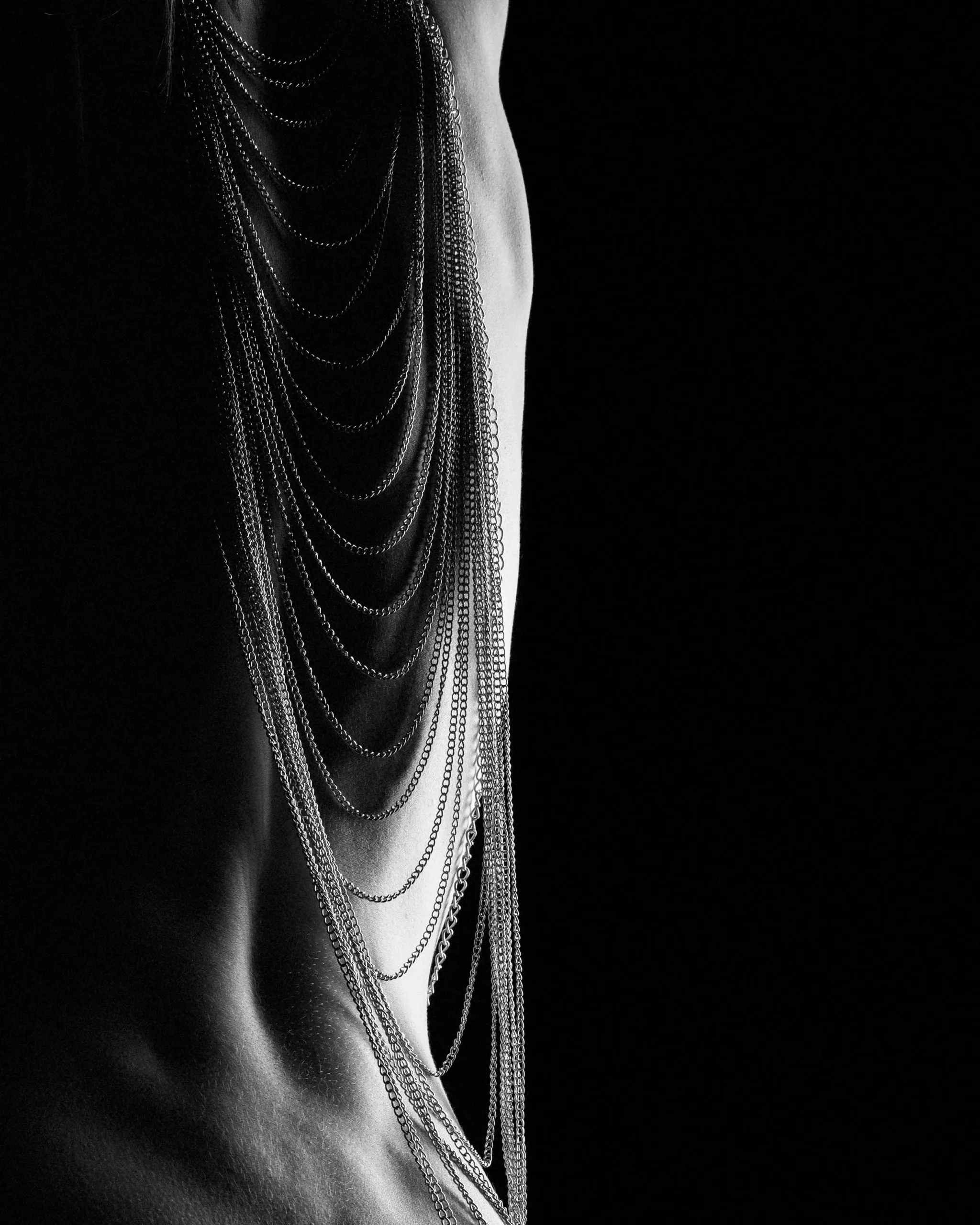 Silver chains curve across a torso emerging from deep black shadow.