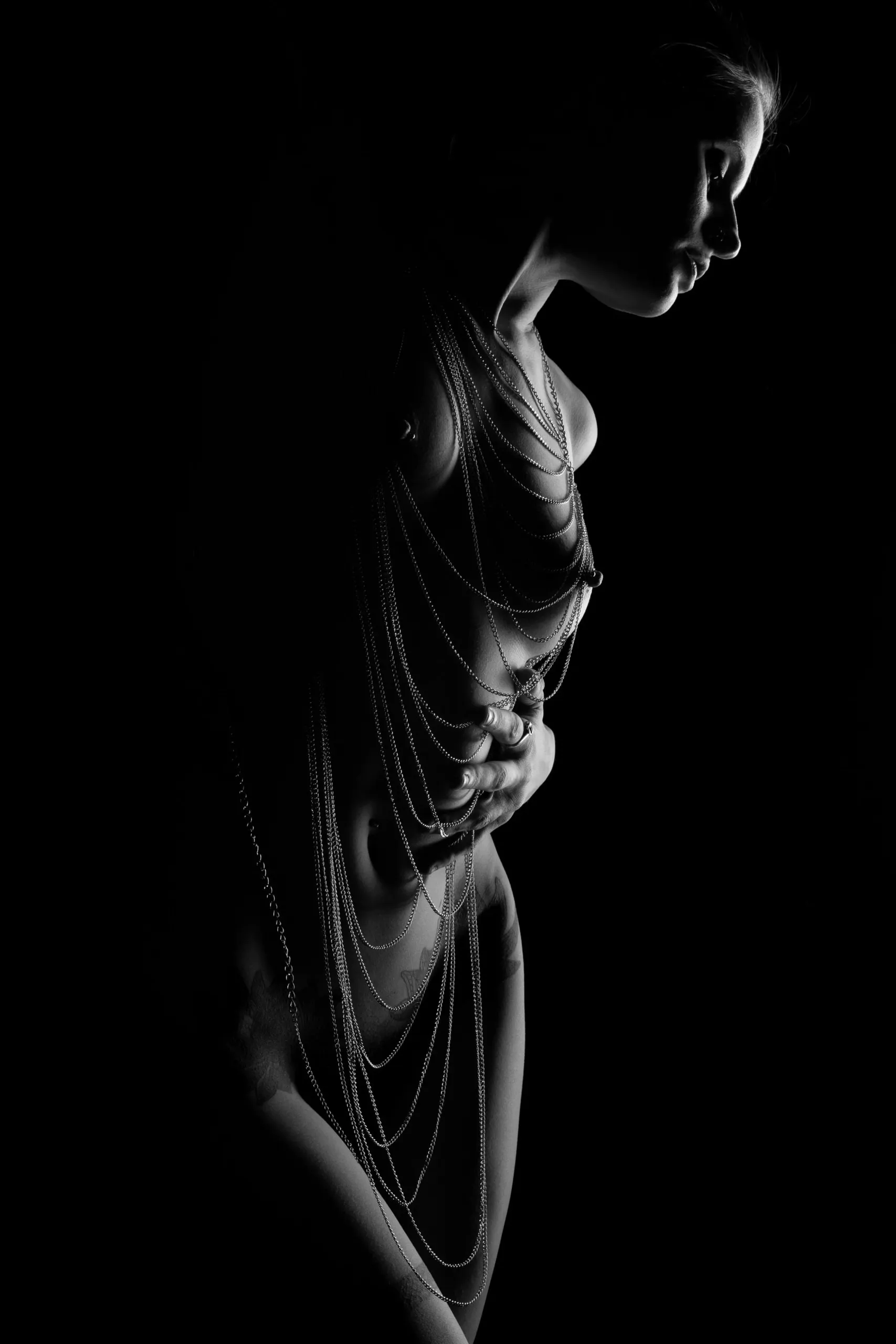 A woman looks down as chains and side light contour her figure.