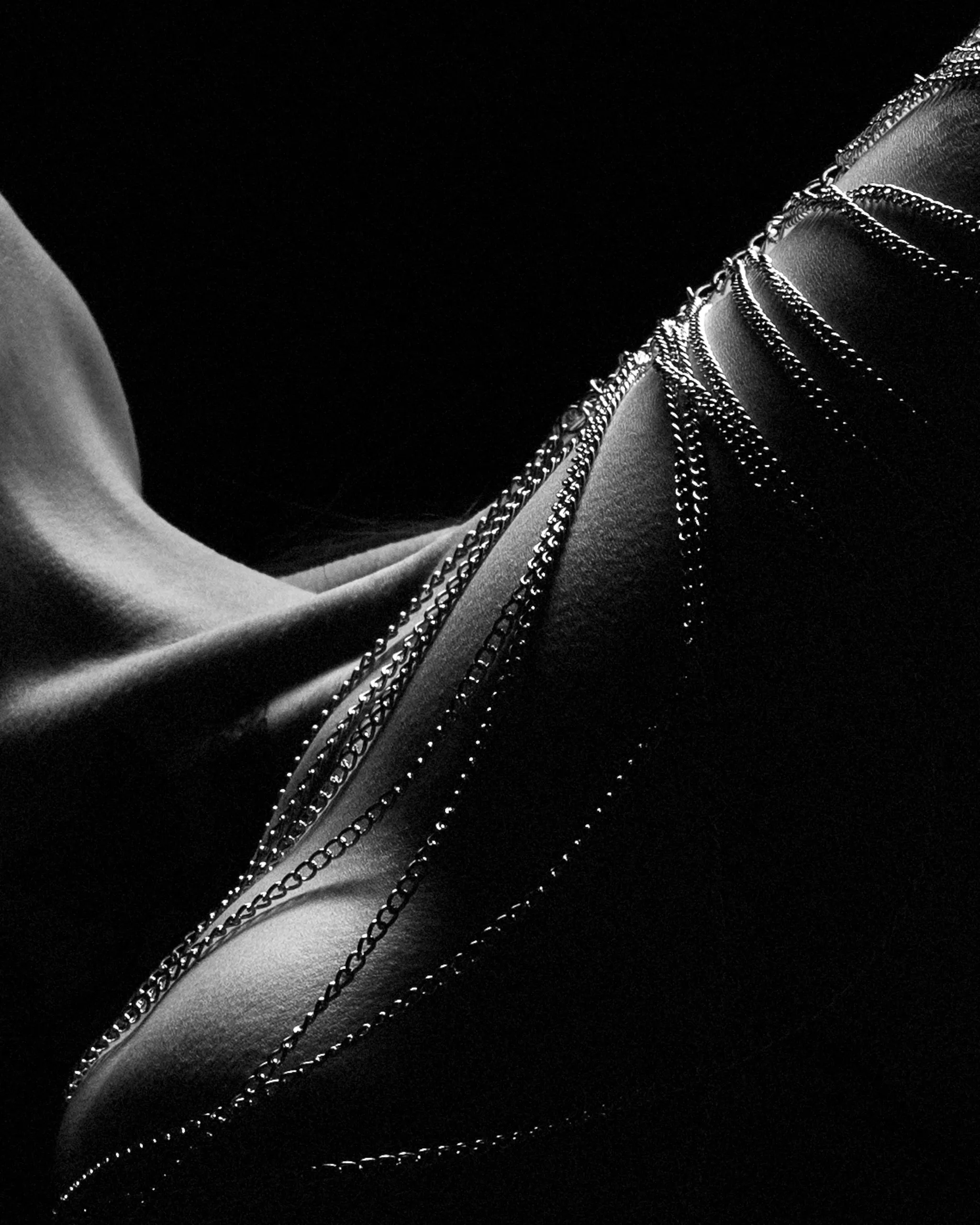A shoulder and torso rise like a landscape beneath strands of chain.