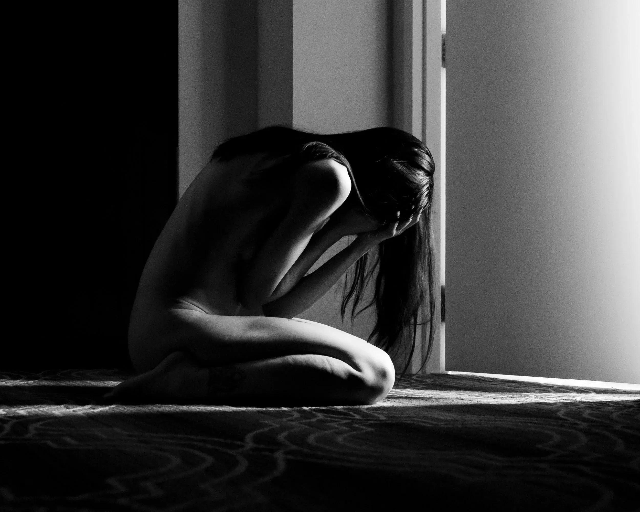 A woman curls on the floor beside a bright doorway in black and white.