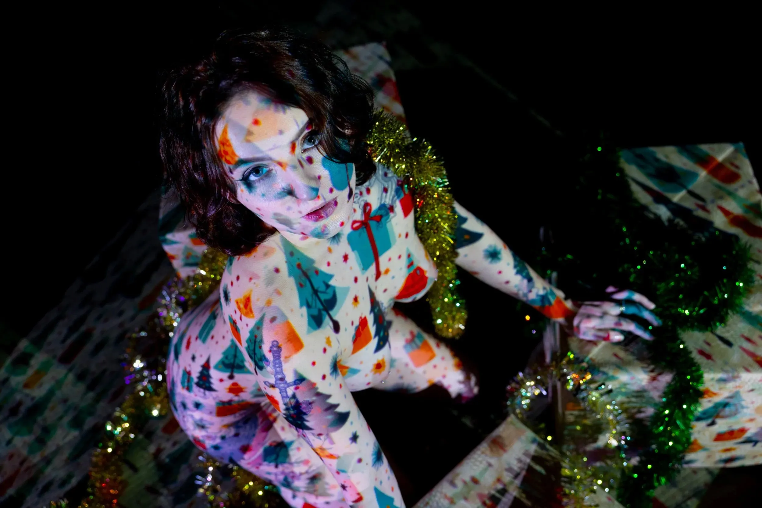 A woman sits among gifts and tinsel beneath multicolored holiday patterns.
