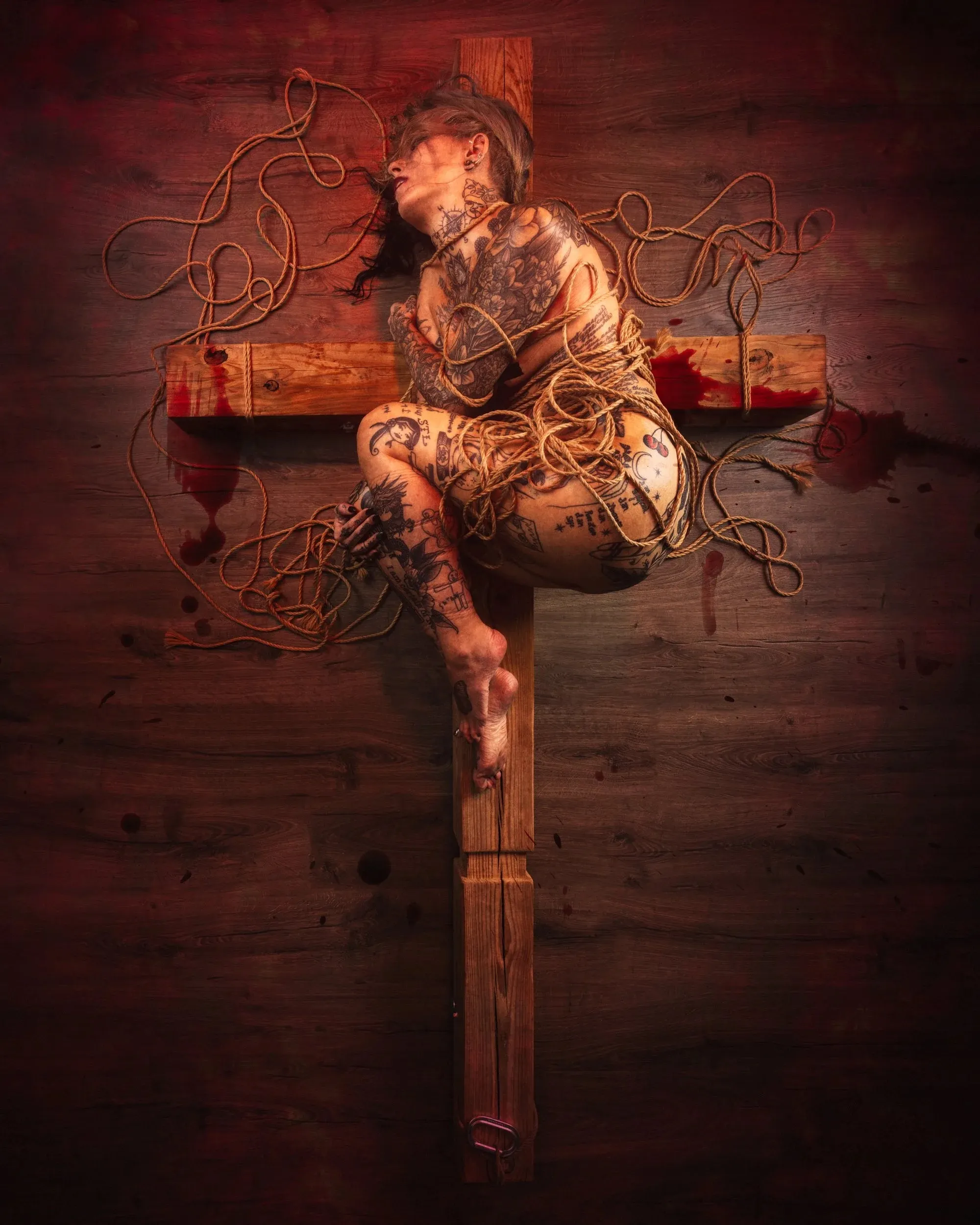 A tattooed figure is bound upright against a full-size wooden cross.