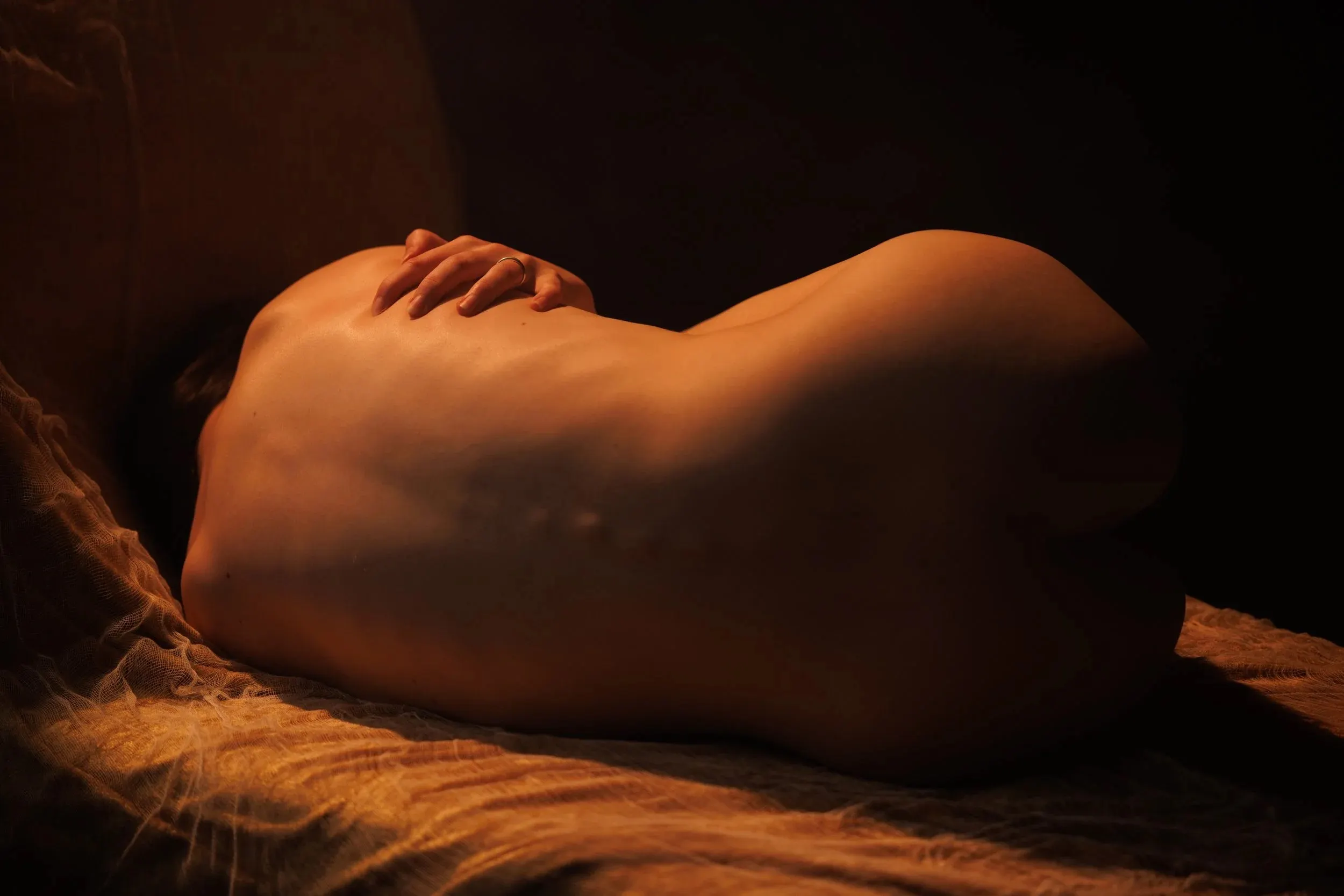 A nude woman is curled up with her back to the camera on textured fabric under soft amber light.