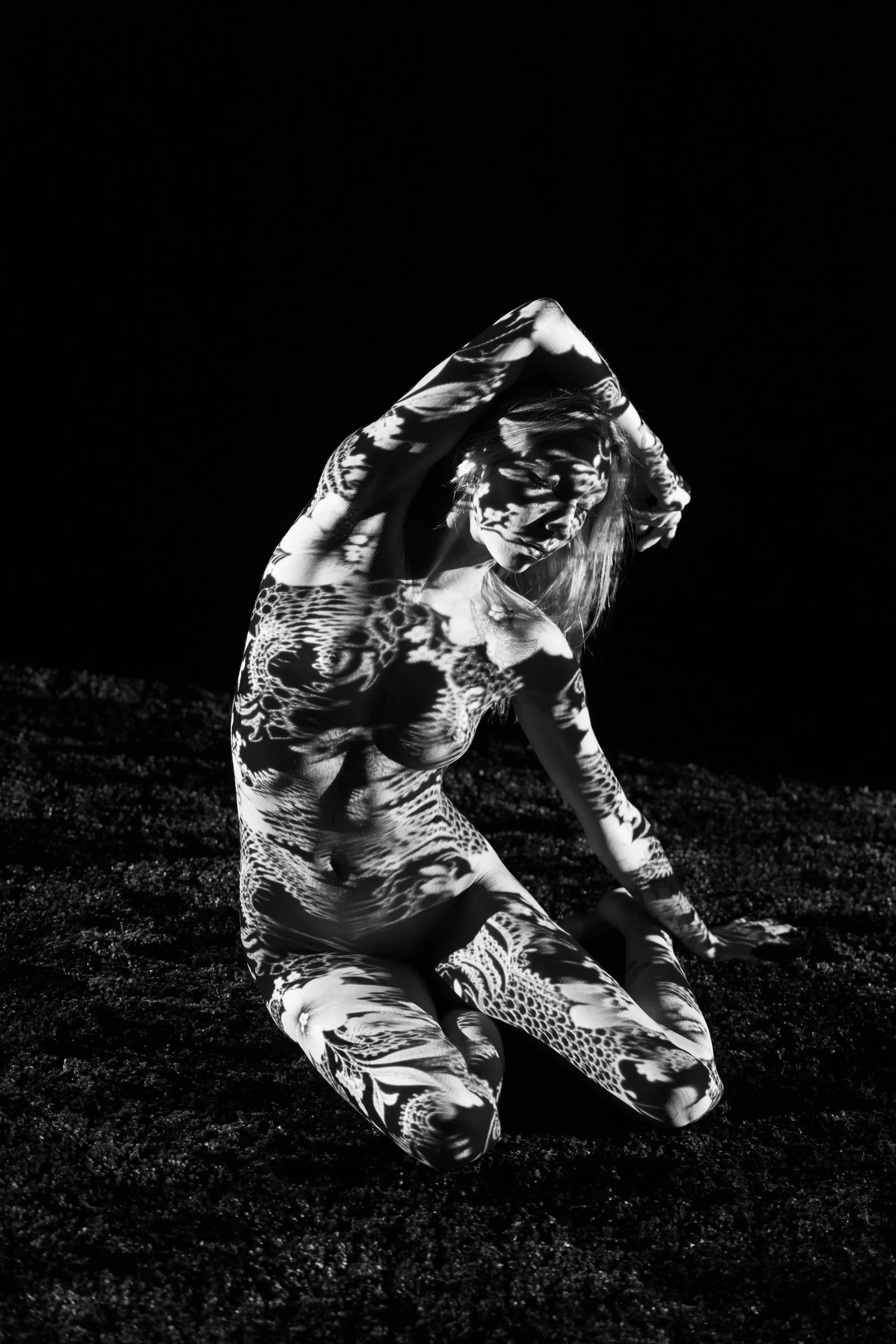 A kneeling woman curves one arm overhead while high-contrast black-and-white floral imagery is projected across her and the floor.