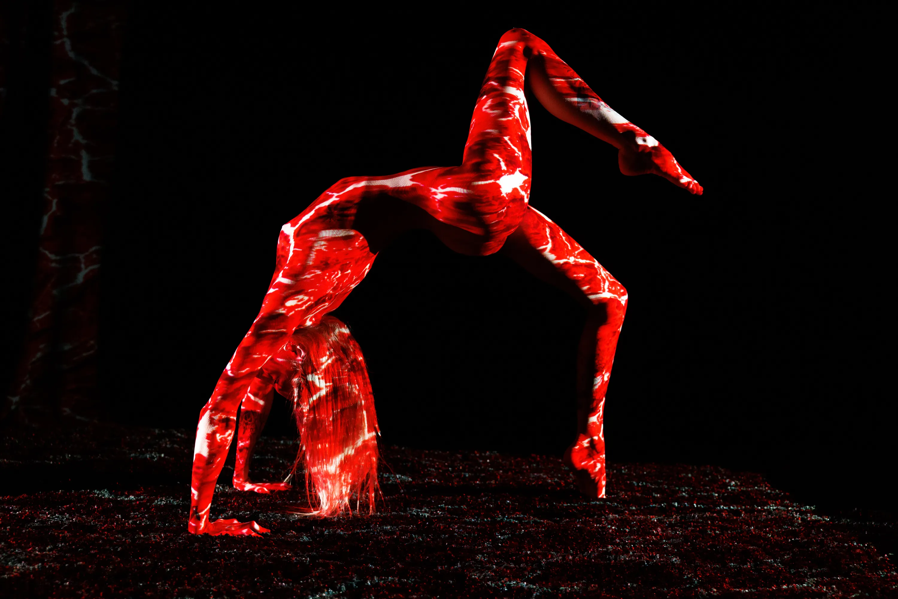 A woman holds a dramatic inverted backbend as vivid red-and-white light patterns sweep across her against black.