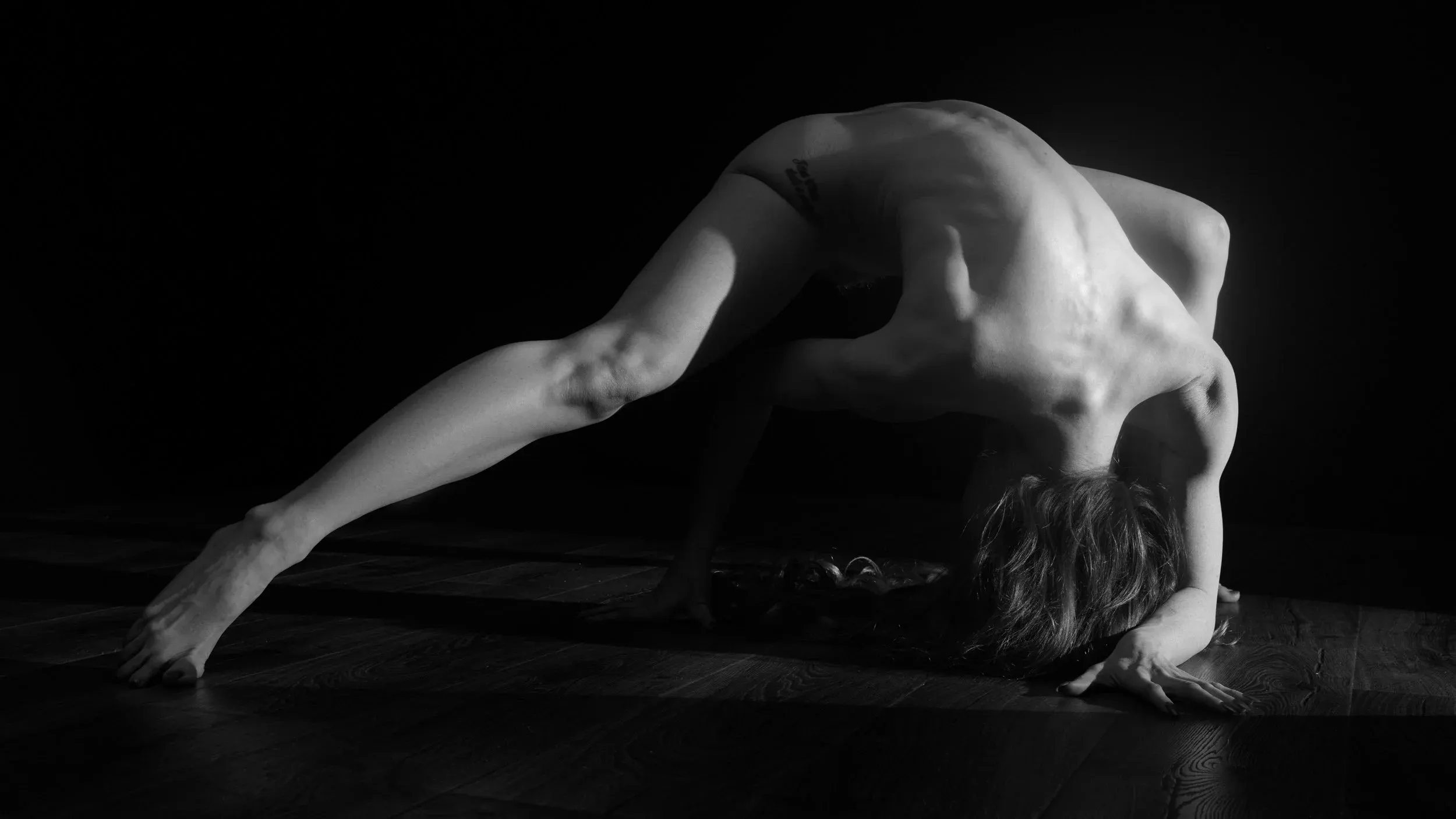 Fine art black-and-white nude photograph of a woman posing on a wooden floor against a black background with hard directional light.