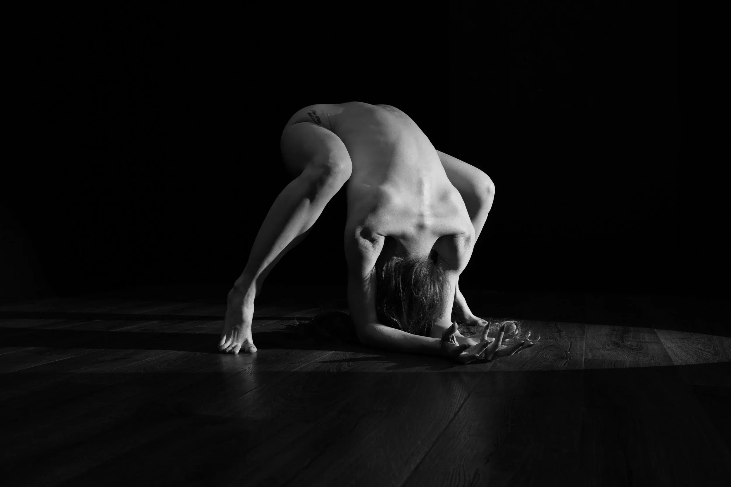 Fine art black-and-white nude photograph of a woman posing on a wooden floor against a black background with hard directional light.