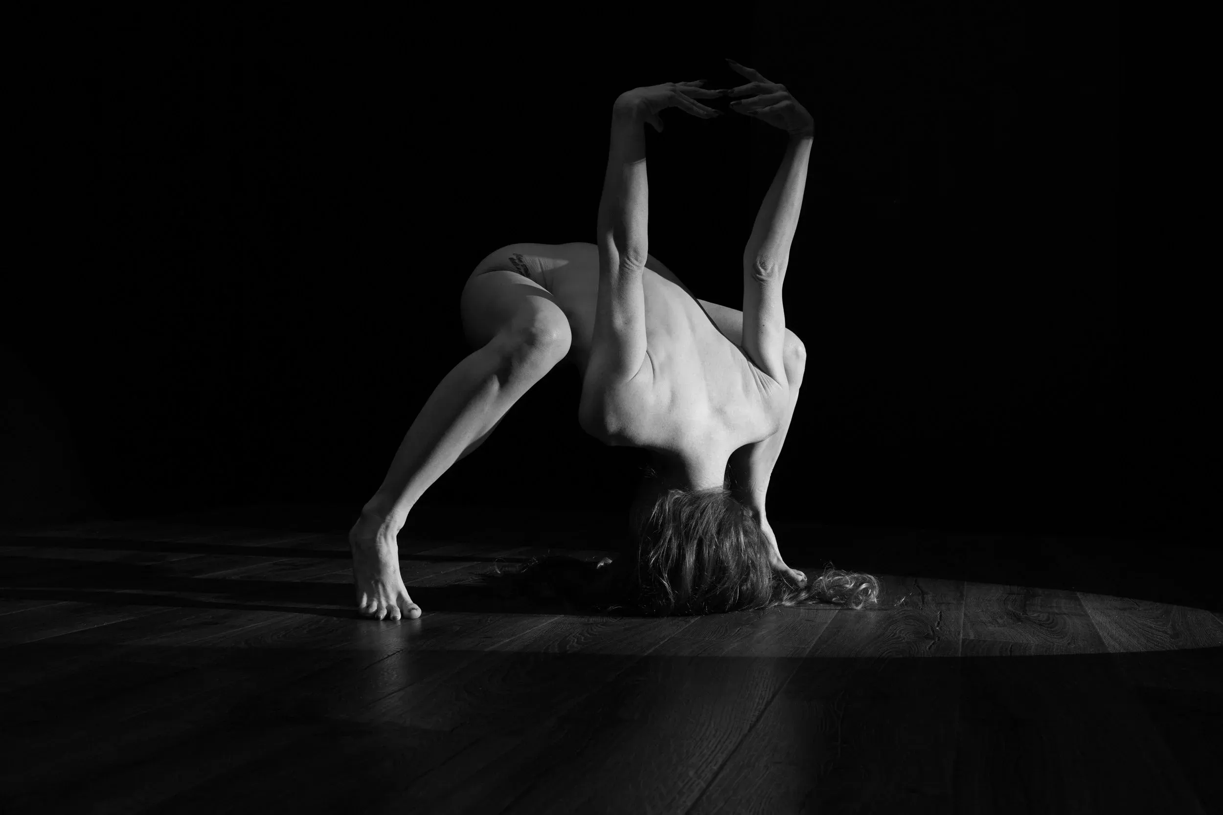 Fine art black-and-white nude photograph of a woman posing on a wooden floor against a black background with hard directional light.