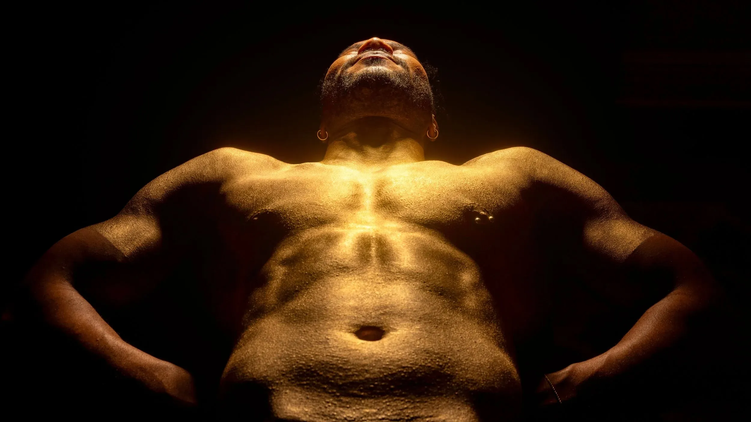 A gold-painted man leans back with hands at his waist against black.