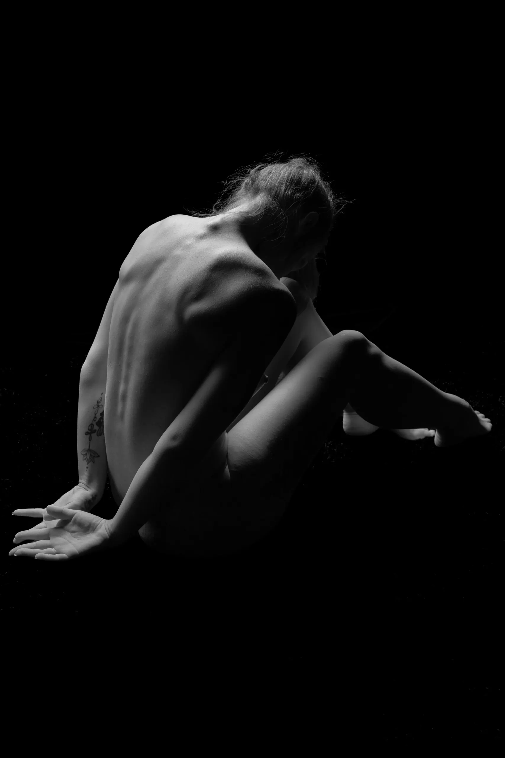 Figurative black and white photograph of a woman who is sitting in a dark space, her back and side to the camera, hands on the floor behind her.