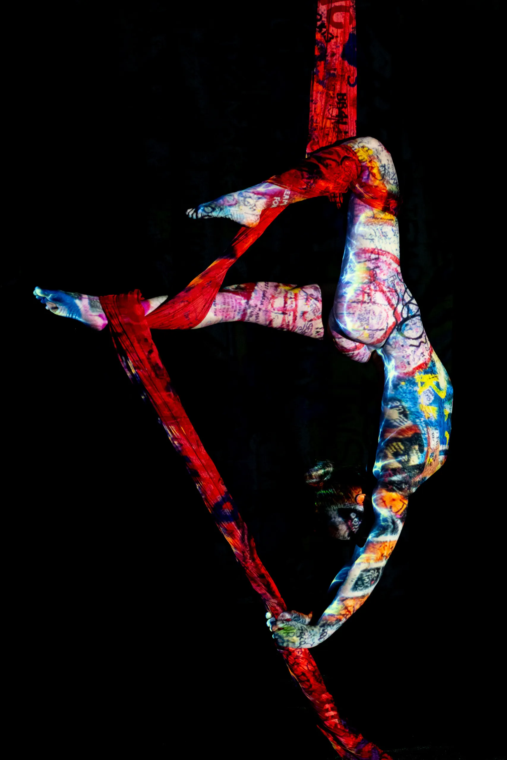 A woman covered in colorful projections hangs inverted from red aerial silks against black.