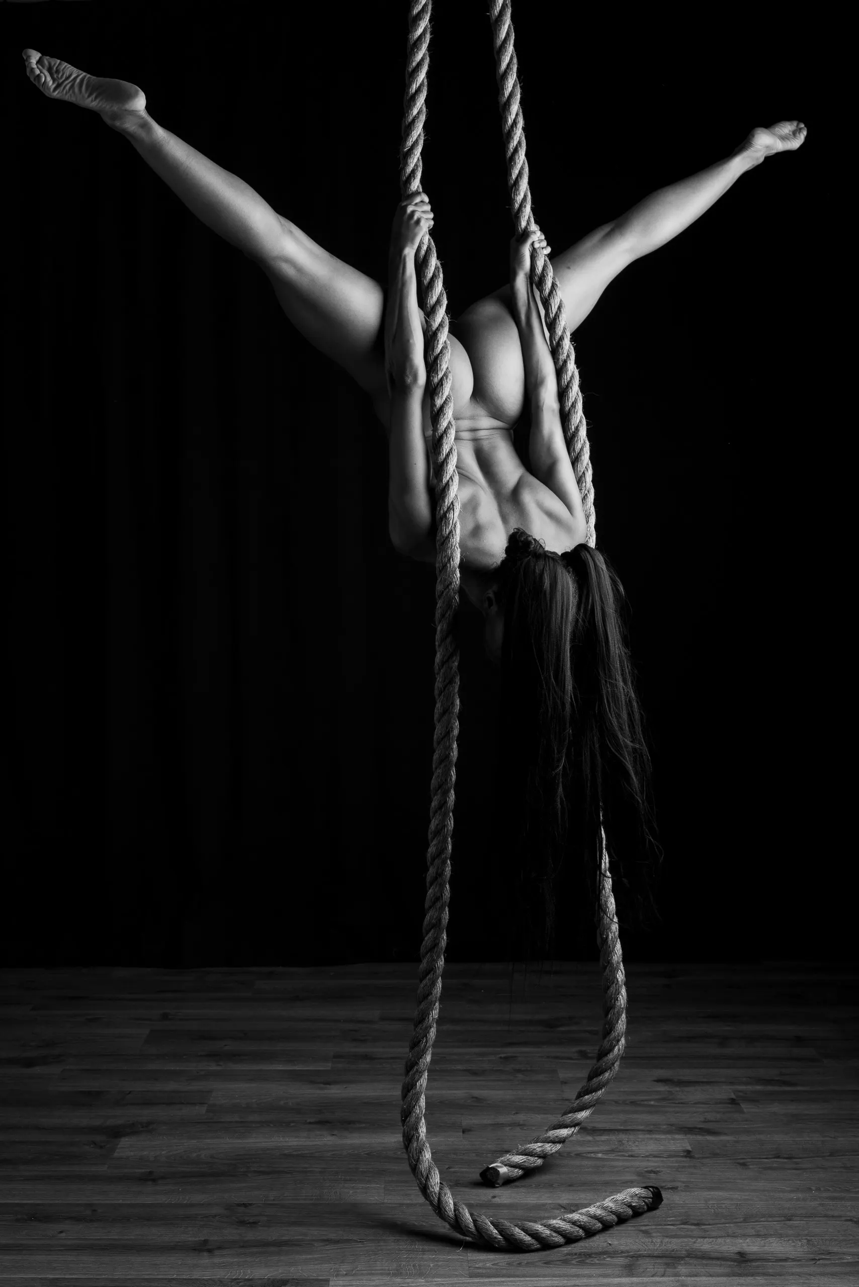 An upside-down woman hangs from a rope above a wooden floor, photographed in black and white.
