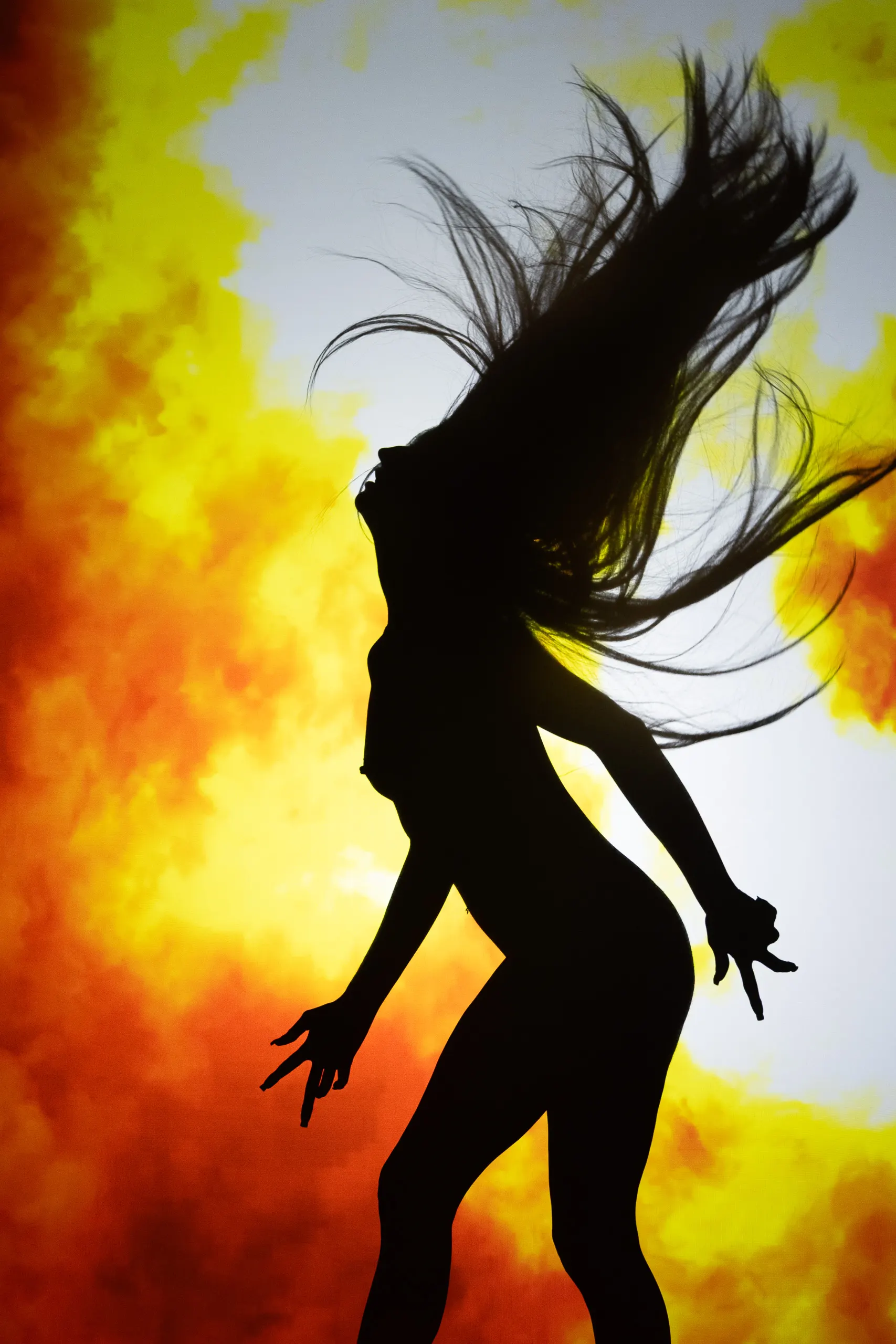 A silhouetted woman throws their hair back against clouds of yellow and orange fire.
