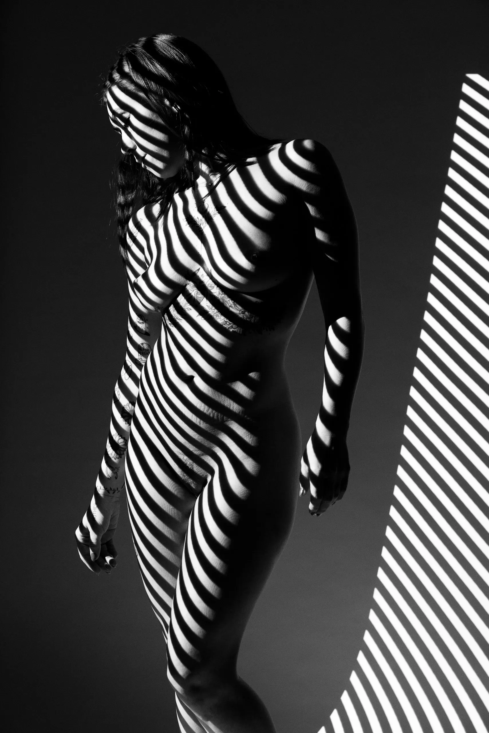Projected white stripes wrap a standing woman as she stands before a white wall, where some of the projected light is also visible.