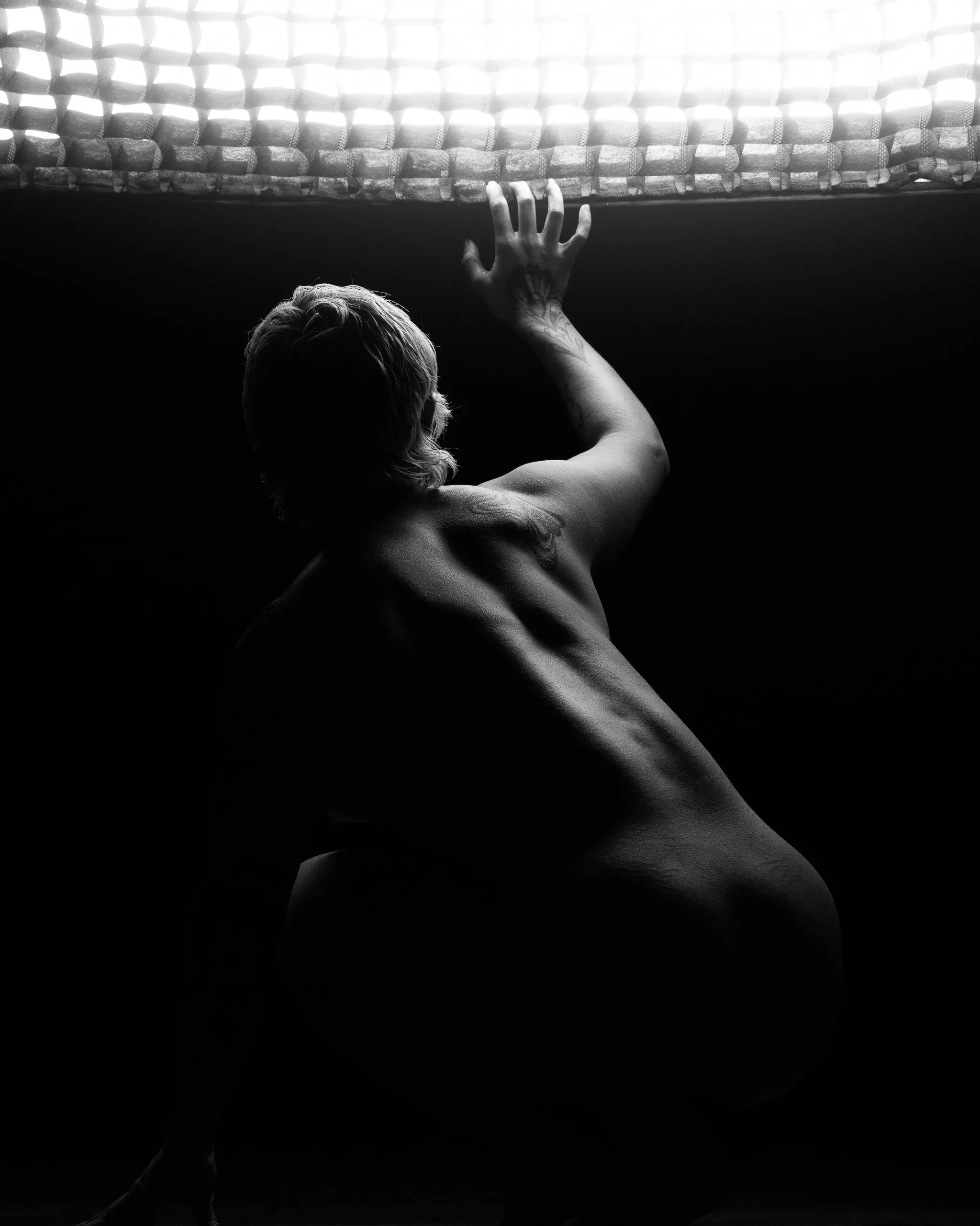 See from behind, a woman reaches toward a bright grid of lights overhead in black and white.