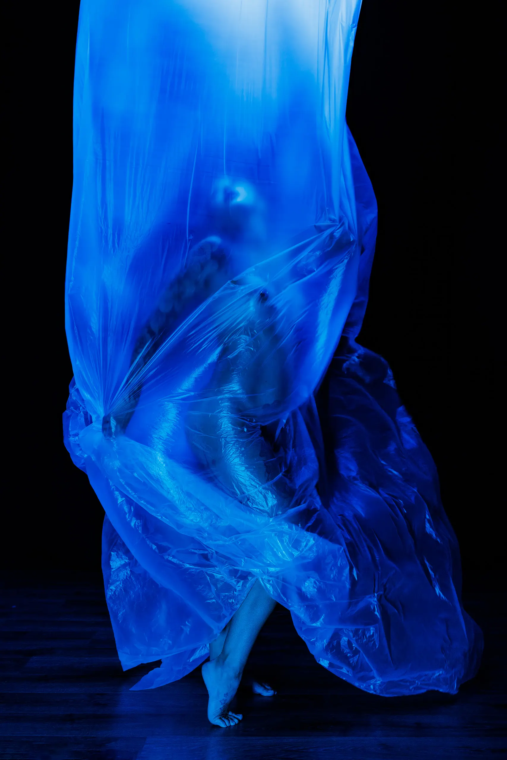 Photo of a woman in a dark studio, surrounded by thin plastic sheets, with blue light shining down from above.