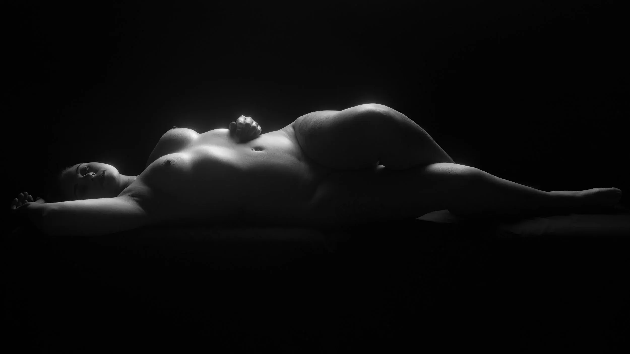 Fine art nude bodyscape of a woman on her back, body turned toward the camera, lit with a single backlight.