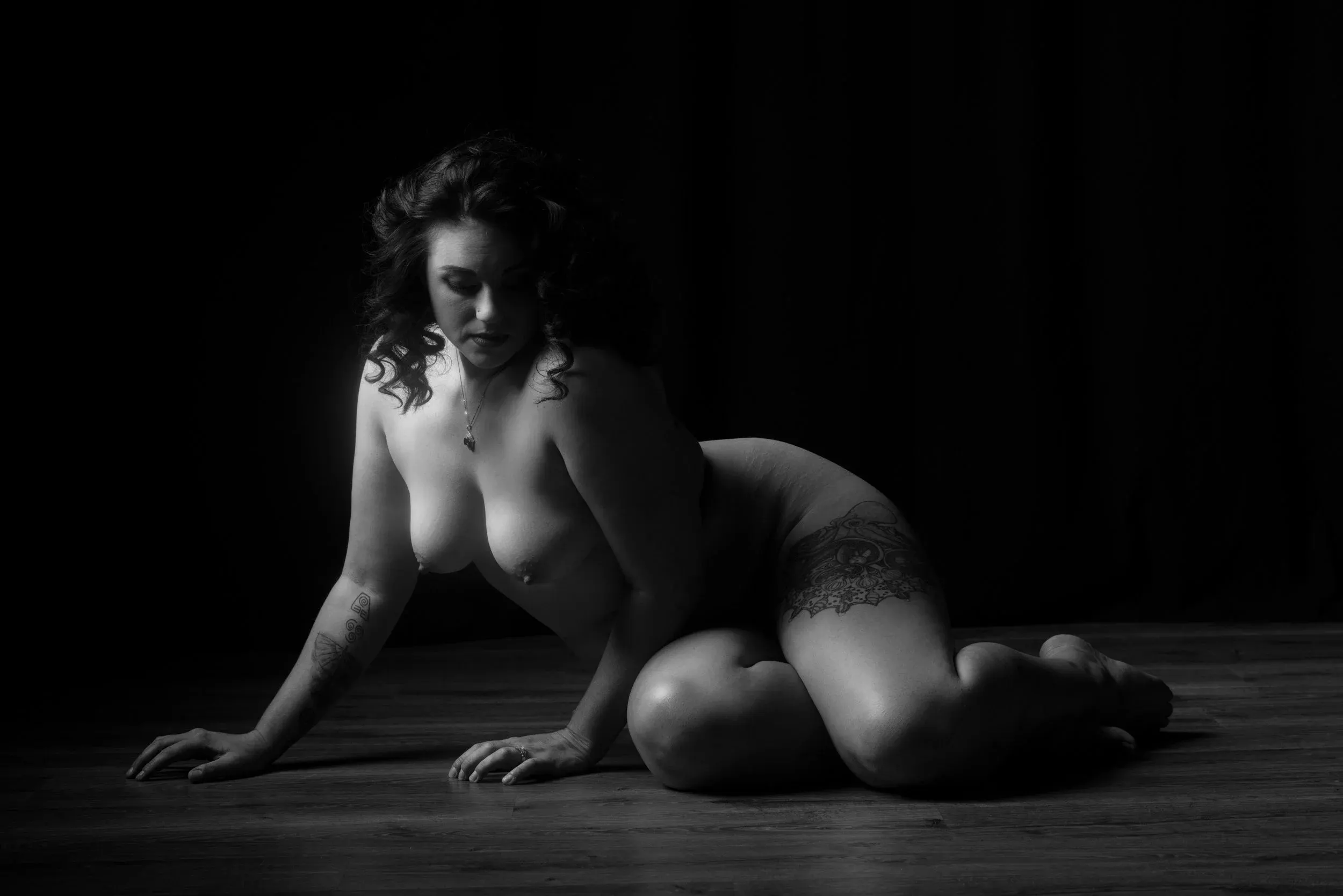 A woman kneels forward on a dark studio floor in side light.