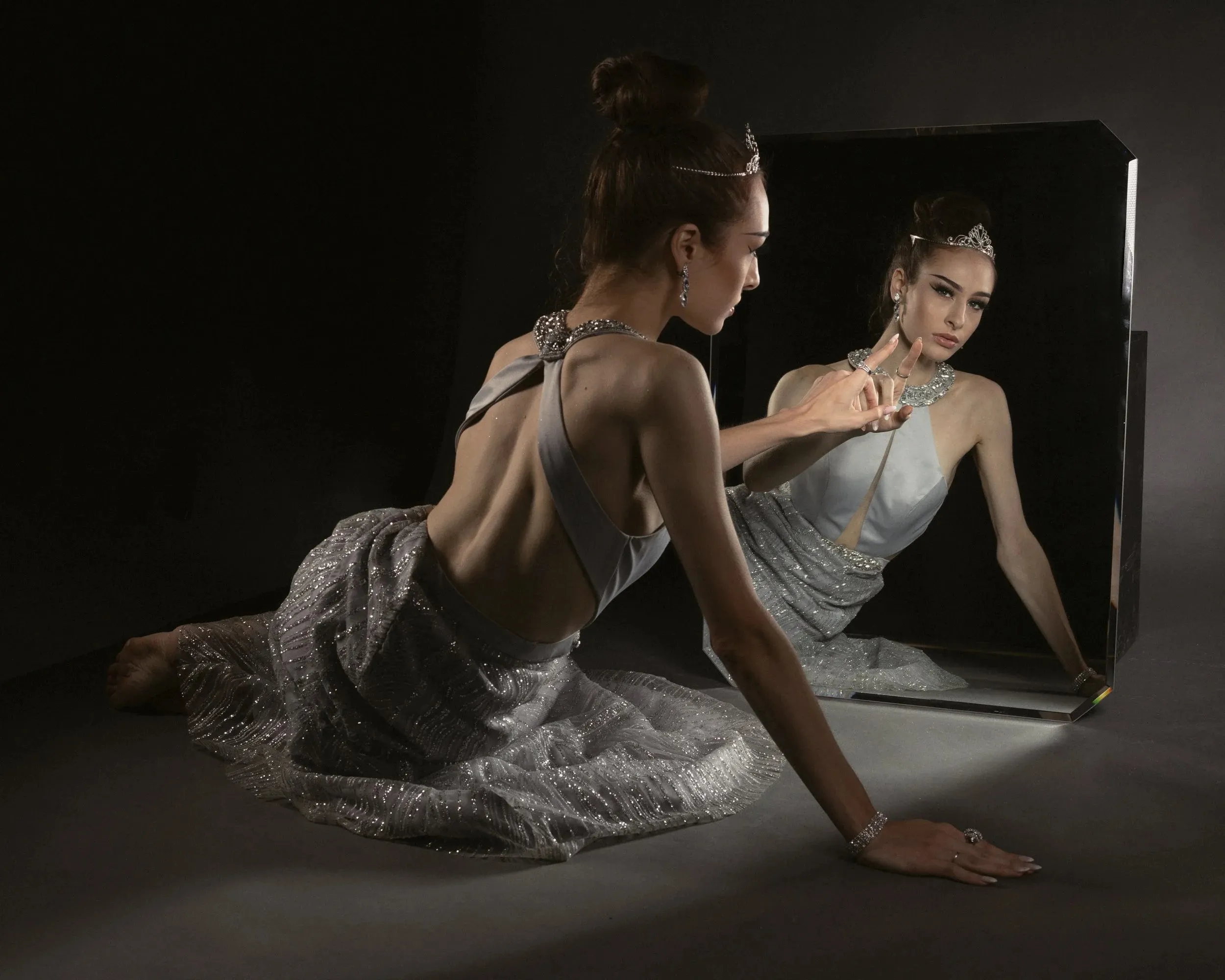 A woman in a silver dress faces her reflection in a large mirror.