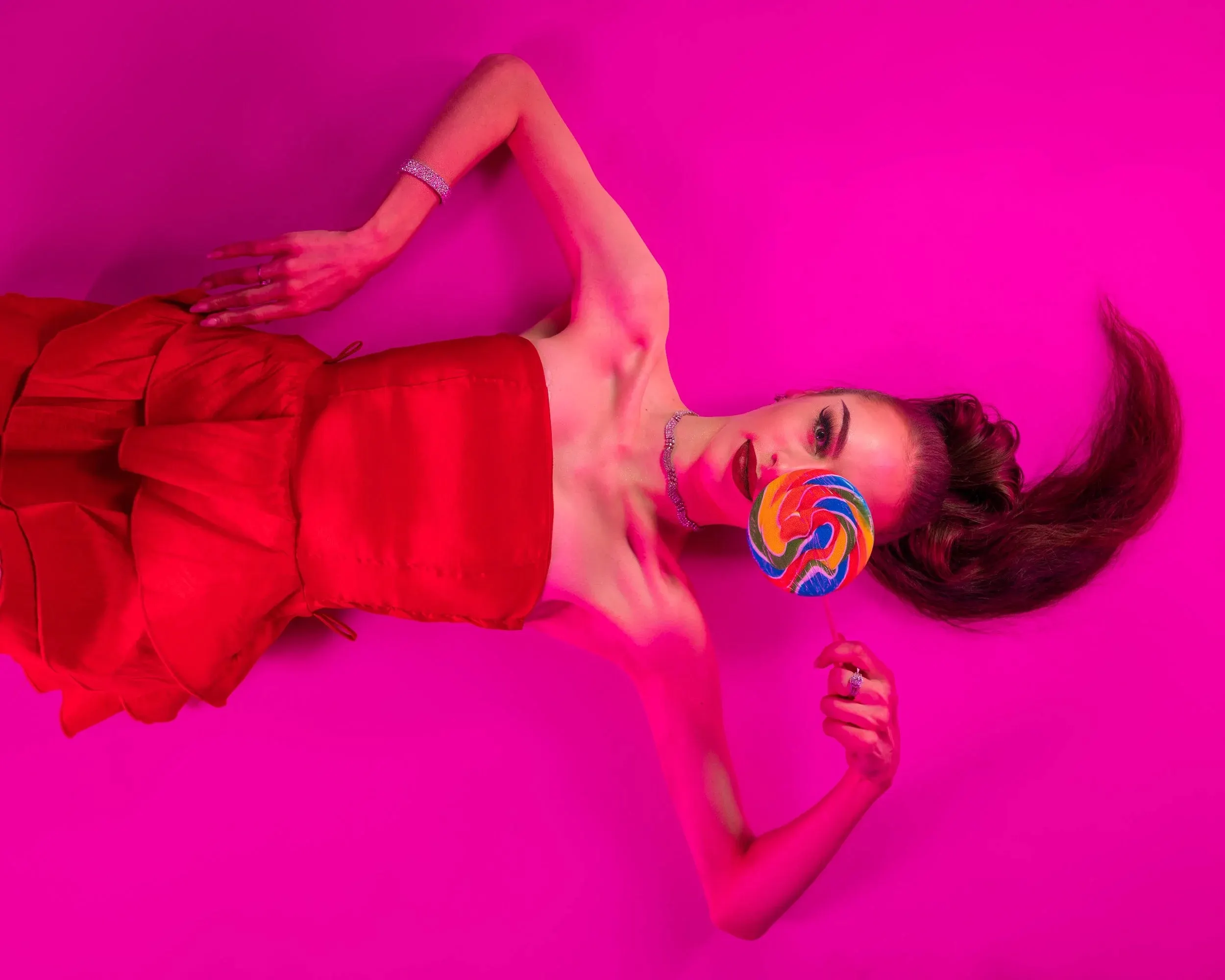 A woman in red lies against a vivid magenta background holding a lollipop.
