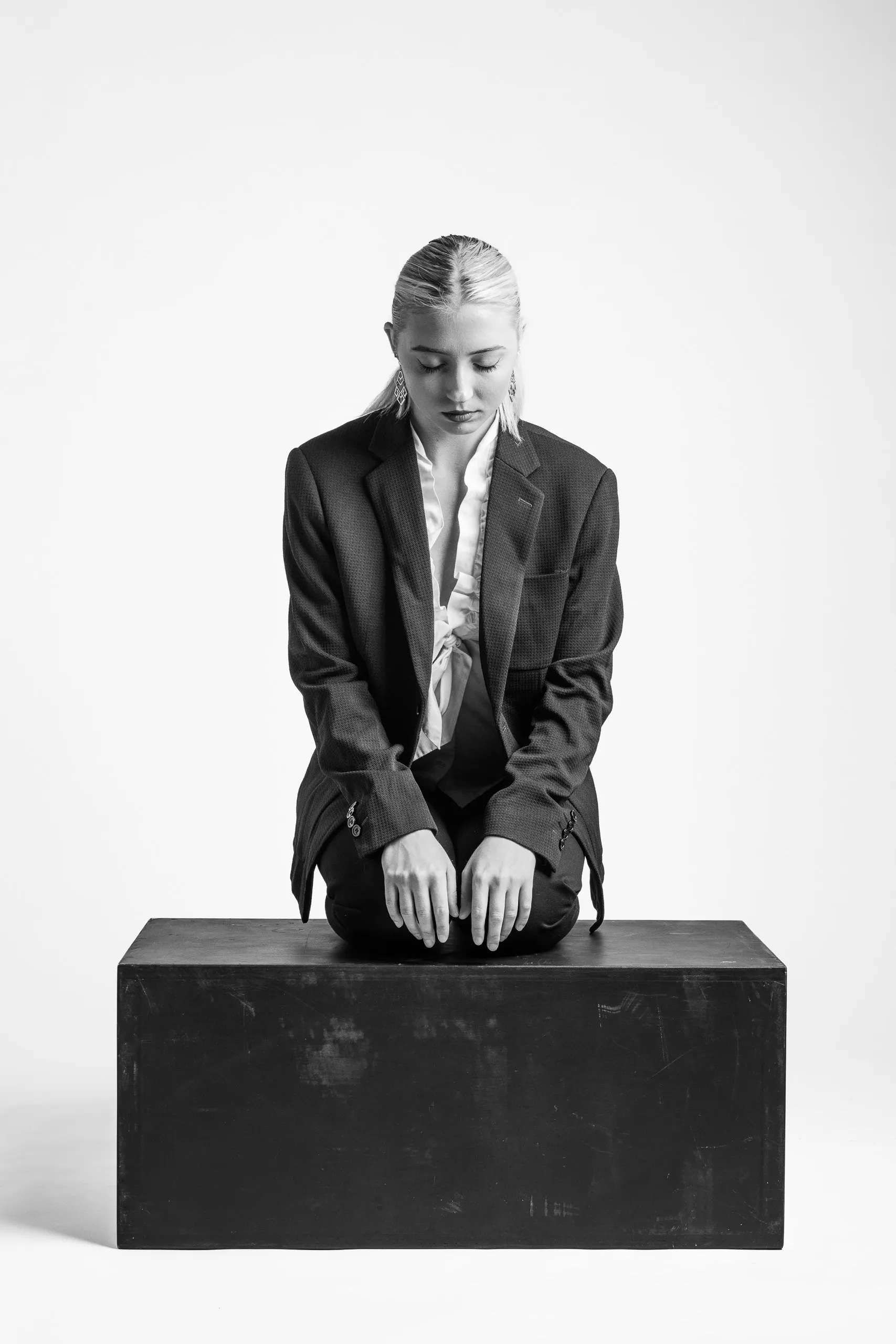 A person with their hair pulled back and wearing a suit jacket is sitting on a box. They are looking down with their hands on their knees.
