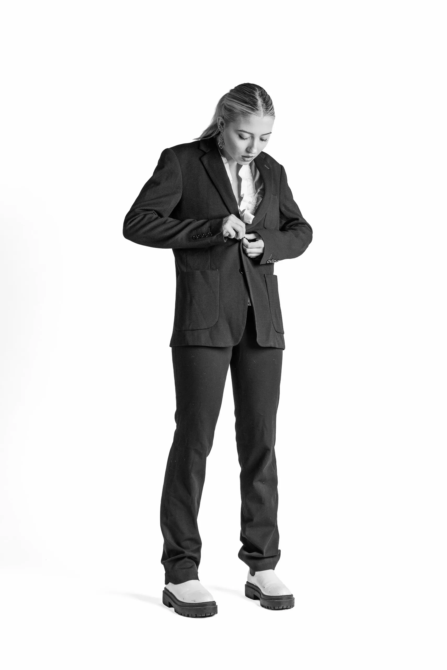 A person wearing a suit stands on a white background, adjusting their jacket. The person has their hair pulled back and is wearing white shoes with dark soles.