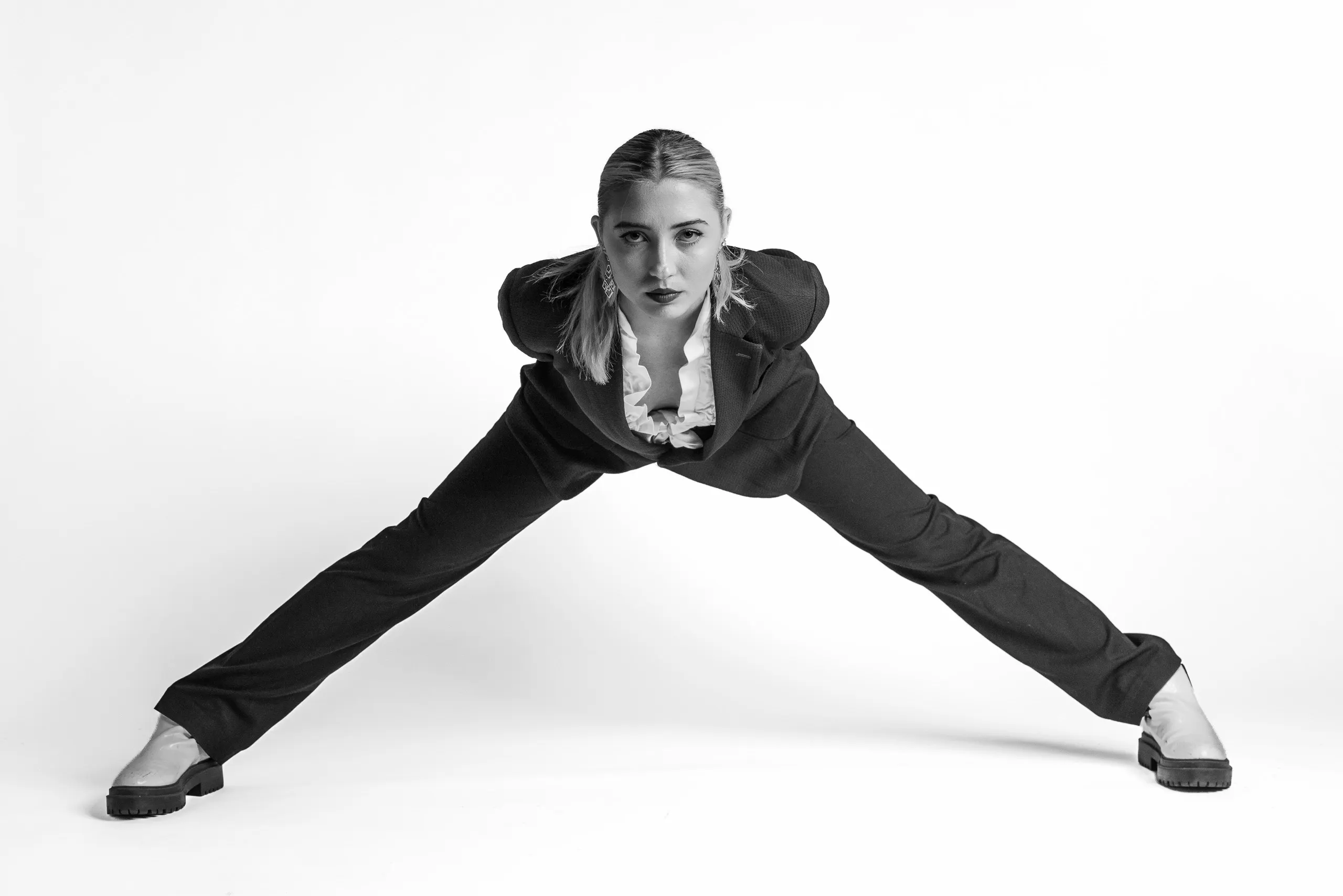 A person with their hair pulled back is performing a split on a white background. They are wearing a dark jacket with a white shirt underneath and white shoes.